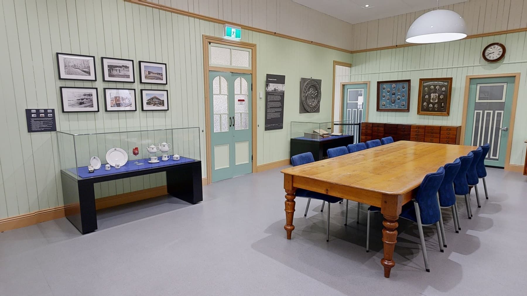 A room with light green panelled walls and a grey floor. On the left, framed photographs are displayed above a glass case containing white crockery on a blue surface. In the centre, a long wooden table with blue chairs runs parallel to the wall. The right side features a window, a wooden plaque, and informational displays. The ceiling is white with recessed lighting and a large round pendant light.