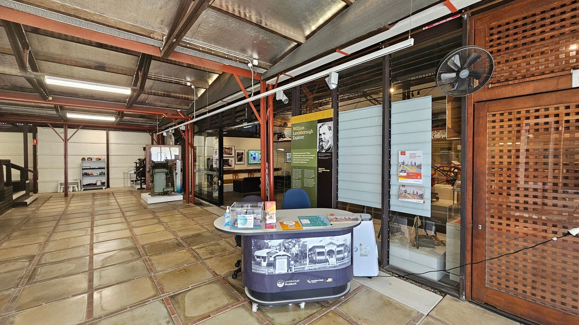 A museum interior with a tiled floor and a metal ceiling. On the right, there's a reception desk with brochures and a chair. Behind it, glass panels display information about William Landsborough. A wall-mounted fan is above. On the left, a display case with various items and a small table with stools are visible. The area is well-lit with fluorescent lights, and the structure features exposed beams.