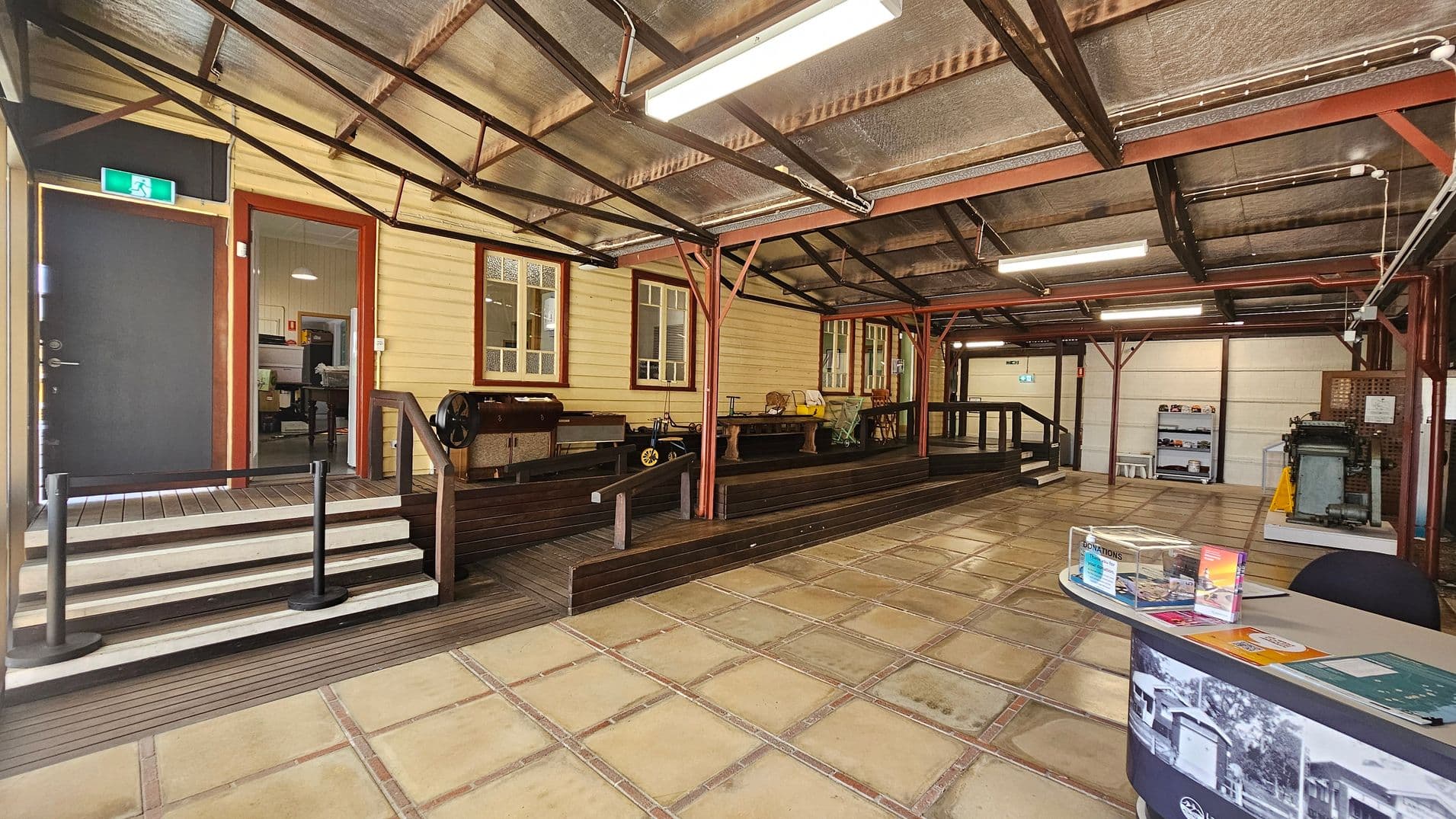 A spacious indoor area with a tiled floor and a corrugated metal ceiling. On the left, there is a black door with an exit sign above it, leading to a room with visible office furniture. Wooden steps and a railing lead up to a platform with vintage machinery and wooden benches. On the right, a curved table displays brochures and a clear box. Shelves with items are visible in the background, along with more machinery. The space is lit by fluorescent lights.