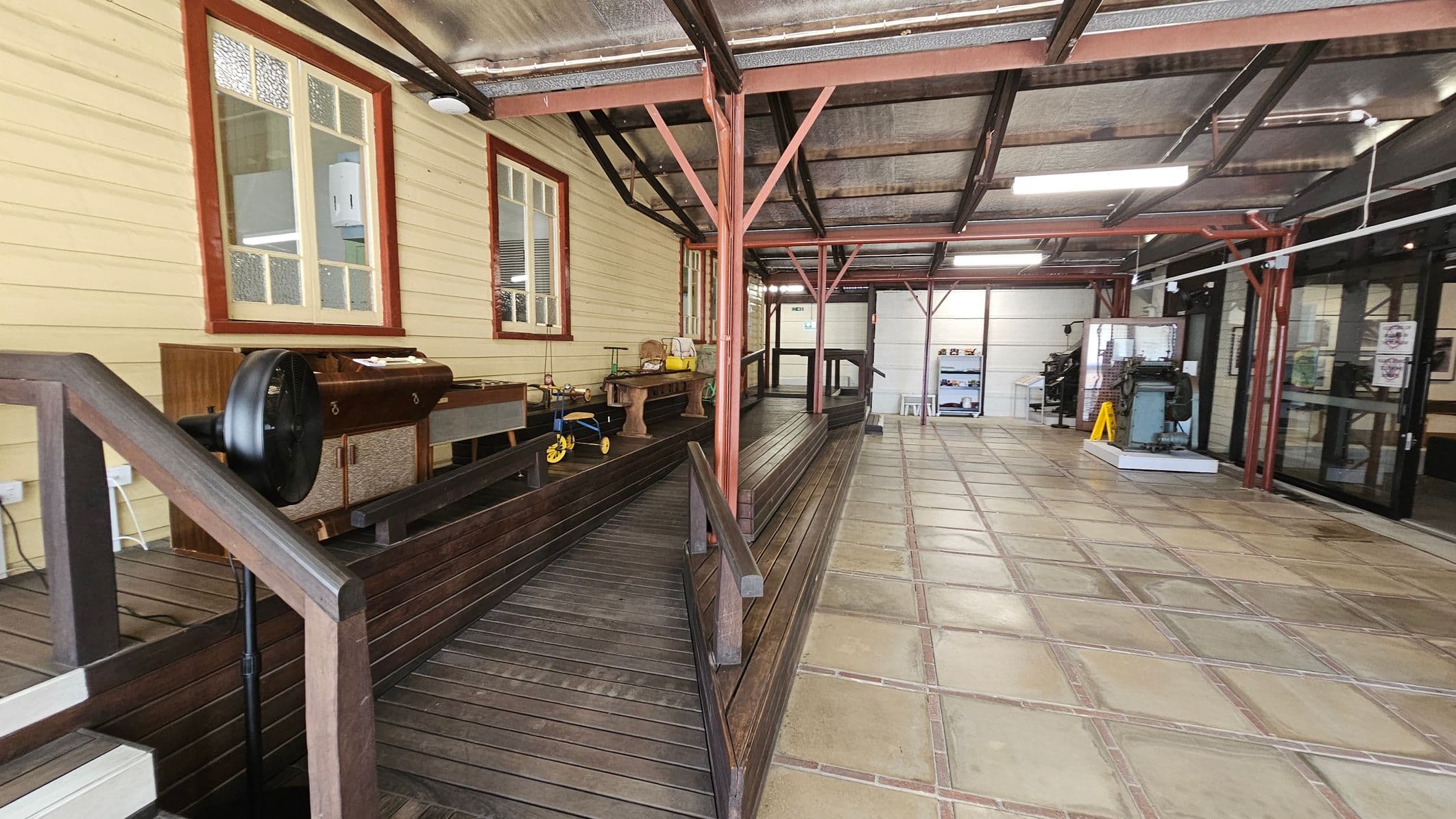 A covered walkway with a wooden ramp on the left, featuring a vintage tricycle and various items on a wooden bench. The wall has two windows with red frames. The right side has a tiled floor and a glass entrance with a yellow caution sign. The ceiling is metal with exposed beams and fluorescent lights. In the background, there is a display case and a machine on a platform.