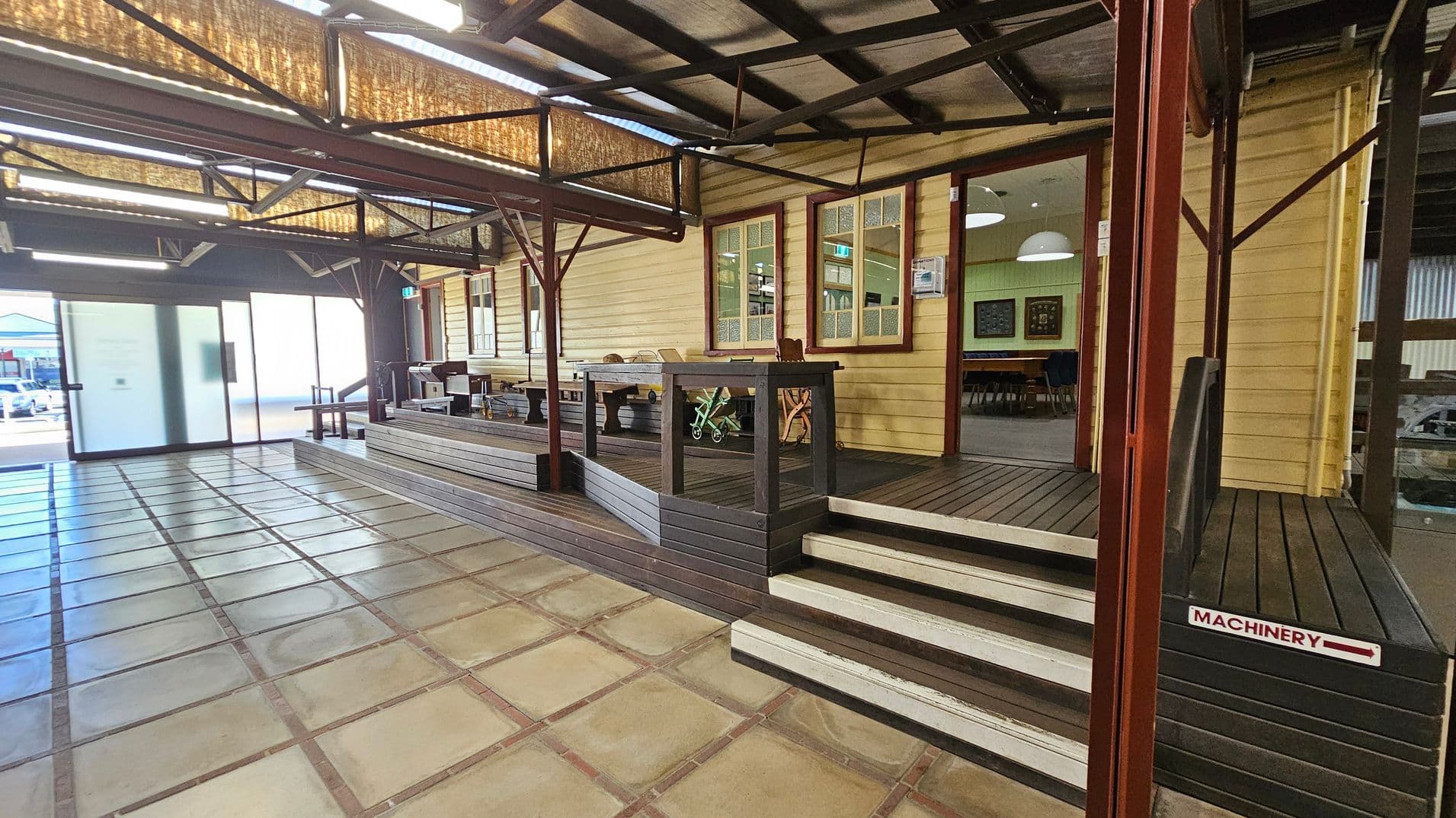 A spacious indoor area with a tiled floor and a wooden decked platform on the right. The platform has steps leading up to a building with cream-coloured wooden walls and large windows. Inside, there are tables and chairs visible. The ceiling has exposed beams and lighting fixtures. On the left, there is a large frosted glass sliding door, and outside, a glimpse of parked cars. A sign on the steps reads "MACHINERY" with an arrow pointing right.