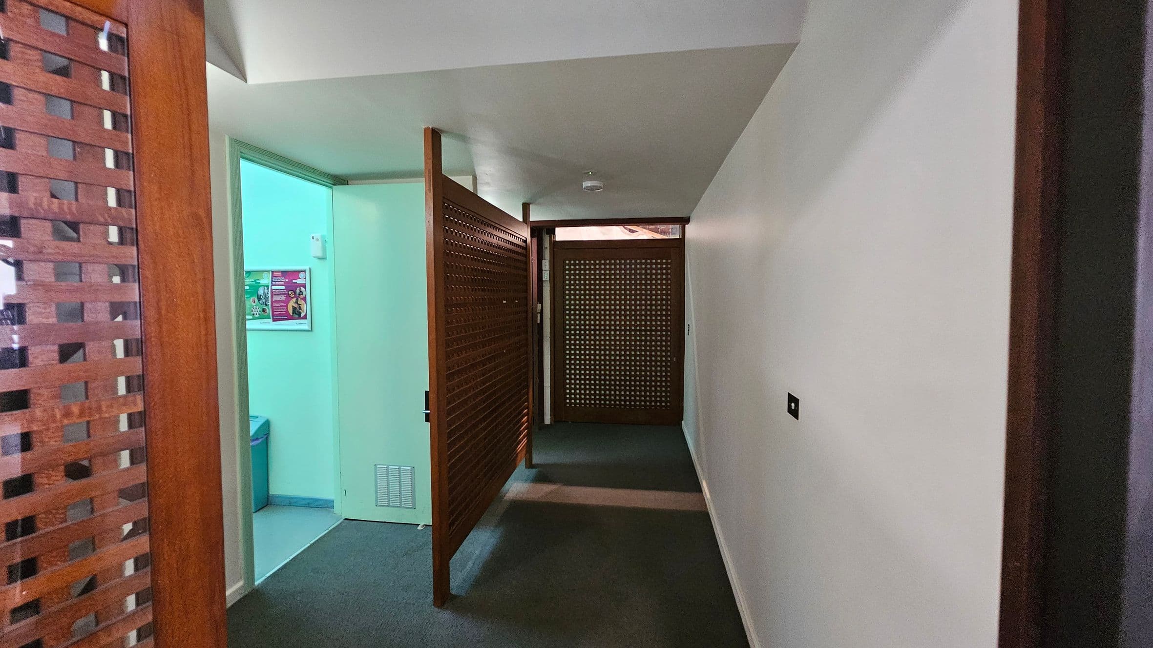A hallway with a grey carpeted floor and white walls. On the left, there's a partially open wooden lattice door leading to a room with a greenish light. Inside, a poster is visible on the wall. Further down the hallway, another wooden lattice door is closed. The ceiling is plain white, and there is a small smoke detector mounted on it.
