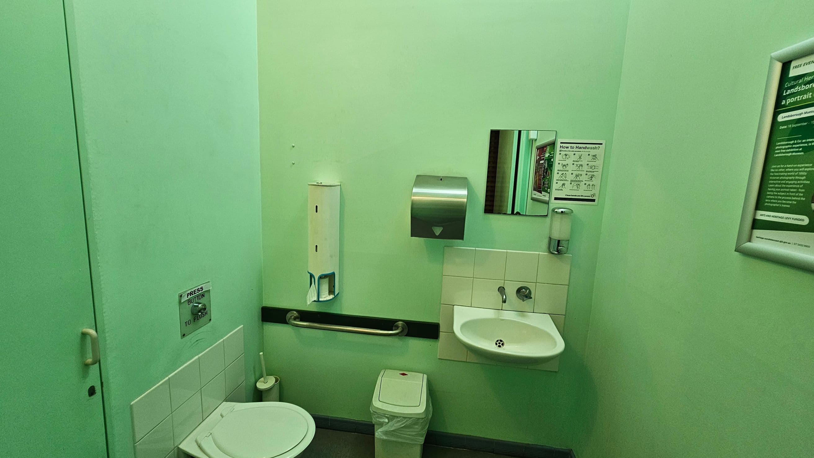 A small bathroom with light green walls. On the left, there's a toilet with a flush button above it and a toilet brush nearby. In the centre, a wall-mounted handrail, a paper towel dispenser, and a small mirror are visible. Below the mirror is a white sink with a soap dispenser. A waste bin is placed under the sink. On the right, there are framed informational posters. The floor is dark, and the ceiling is not visible.