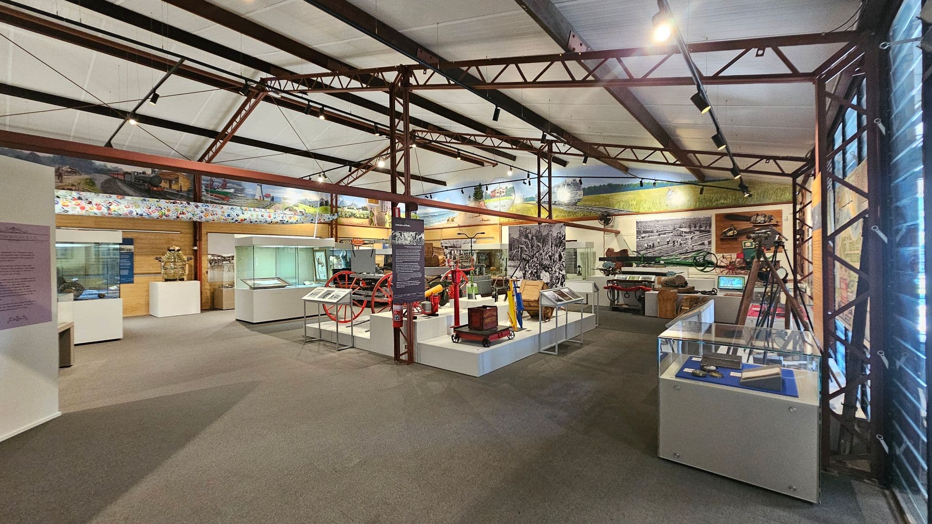 A museum interior with various exhibits displayed in glass cases and on platforms. On the left, there are informational panels and display cases. In the centre, historical machinery and artefacts are showcased, including a red wheel and other equipment. The right side features more display cases and a tripod. The ceiling has exposed metal beams and track lighting, and the floor is carpeted. Large murals and photographs decorate the walls.