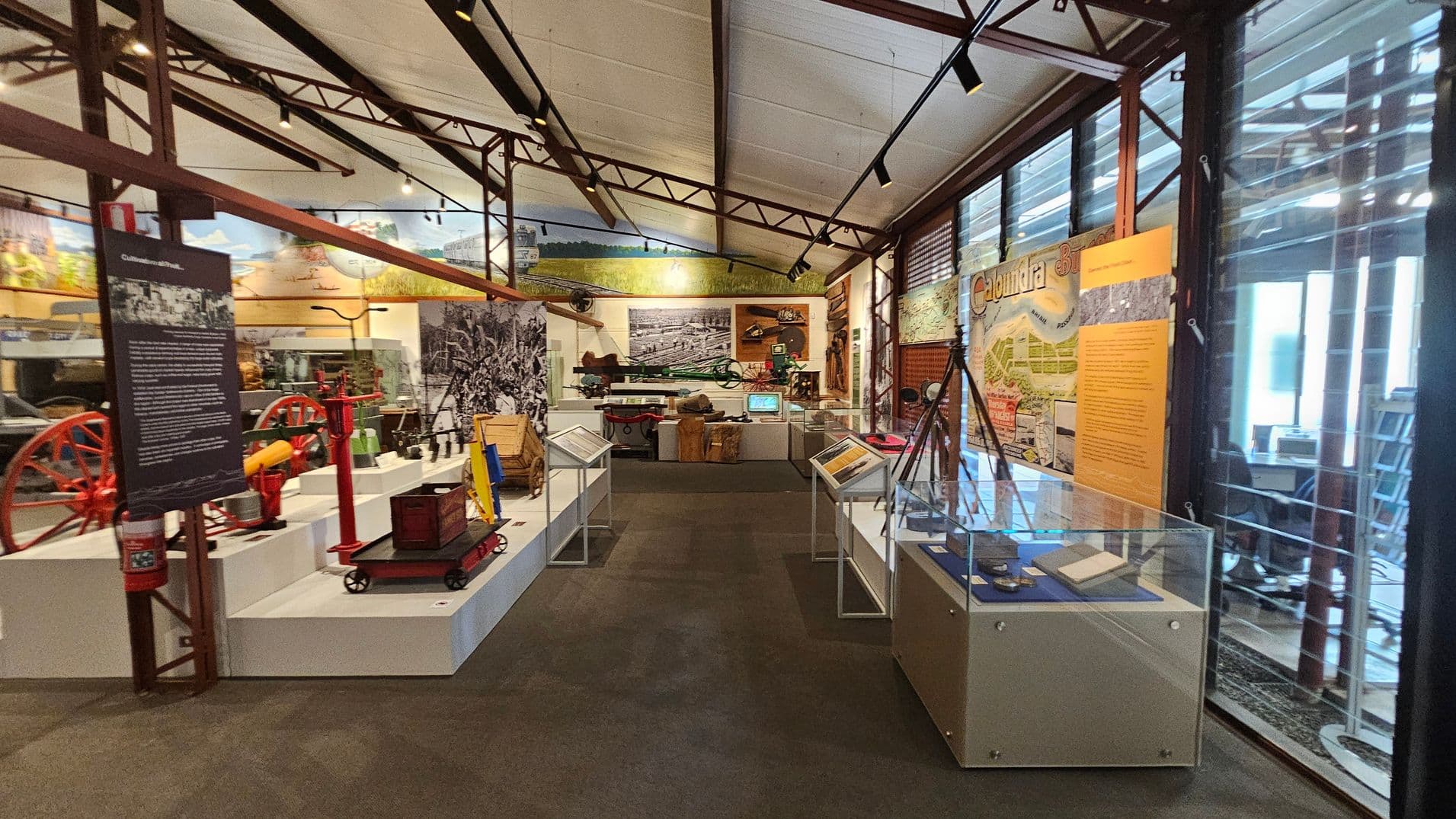 A museum interior with various exhibits. On the left, there are red and yellow machinery parts displayed on white platforms. In the centre, a large black-and-white photograph is mounted on the wall, with more machinery and artefacts below it. On the right, glass display cases contain documents and small items. The walls feature informational panels and historical images. The ceiling is supported by metal beams, and the floor is carpeted. Large windows on the right allow natural light in.