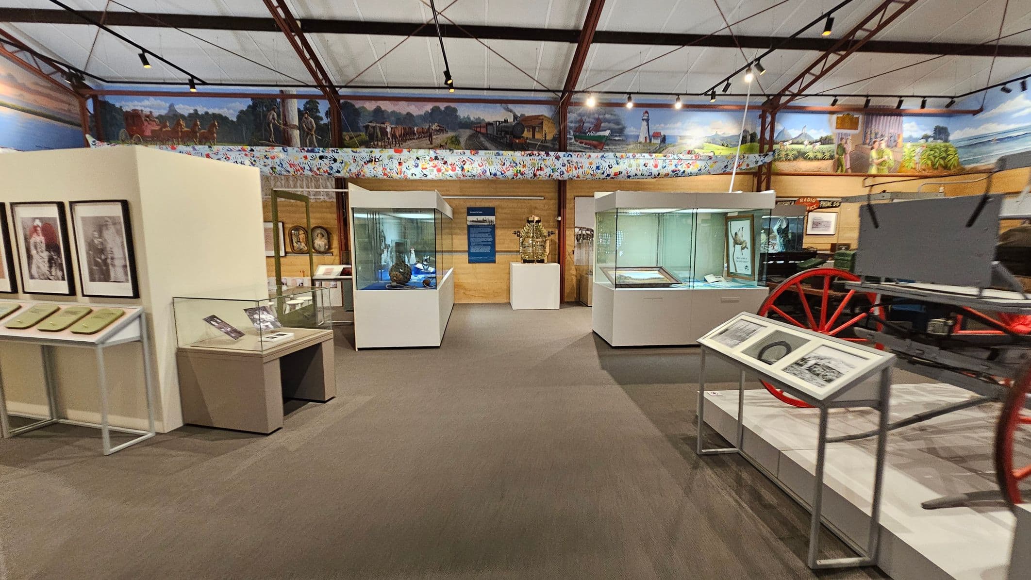 A museum interior with display cases and exhibits. On the left, there are framed photographs and documents on a stand. In the centre, glass display cases contain various artefacts, with a wooden wall and a blue information panel behind them. On the right, a red-wheeled carriage is partially visible, accompanied by informational panels. The ceiling is supported by metal beams, and a mural runs along the upper walls, depicting historical scenes. The floor is carpeted.