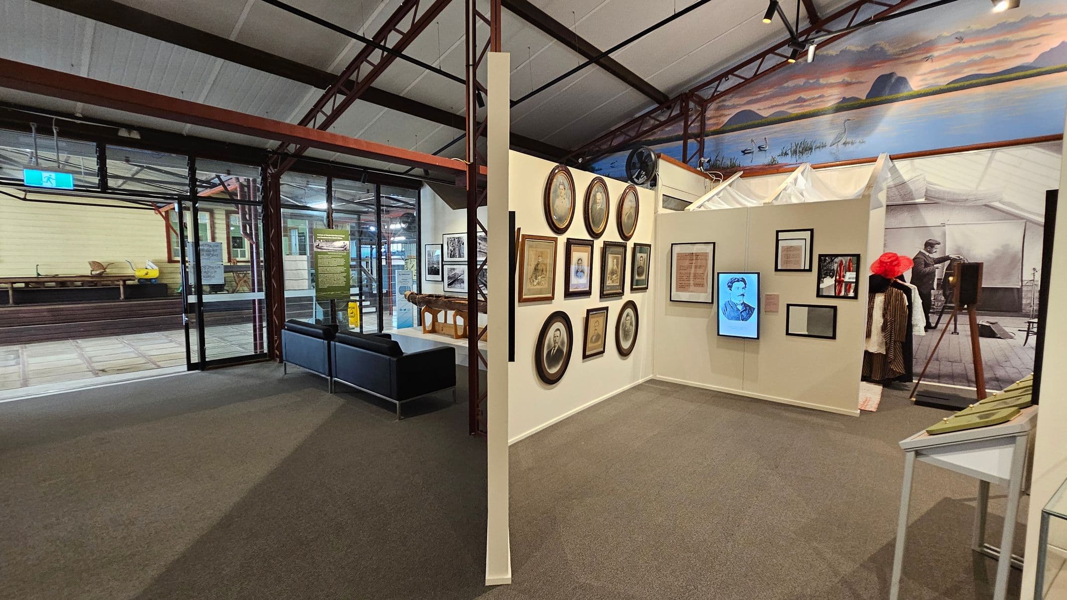 A museum interior with a carpeted floor and high ceiling. On the left, there's a glass entrance with a view of an outdoor area featuring a yellow building and wooden benches. In the centre, a wall displays various framed portraits and documents. To the right, there's a historical exhibit with vintage clothing and an old camera. A mural of a landscape with water and hills is visible above the exhibits. A small seating area with a black sofa is near the entrance.