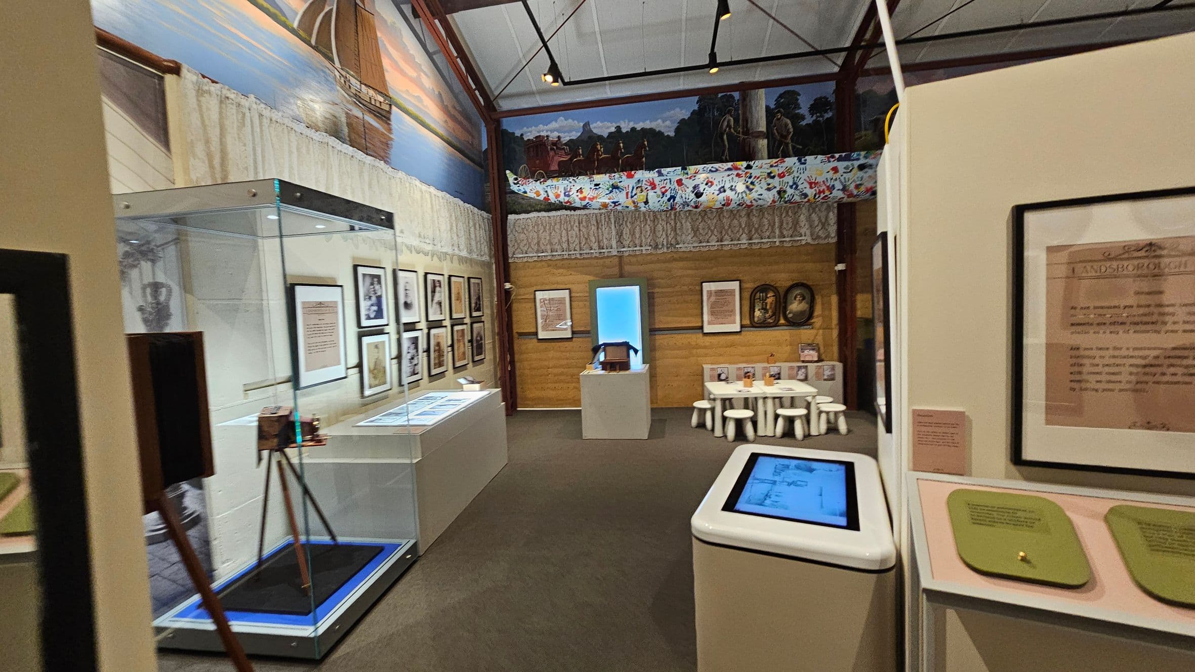 A museum exhibit with framed documents and photographs on the walls. On the left, a glass display case contains an object on a tripod. In the centre, a small table with white stools is set up, possibly for interactive activities. The background features a mural with a canoe and handprints. The ceiling is high with visible beams, and the floor is carpeted. An interactive screen is on the right.