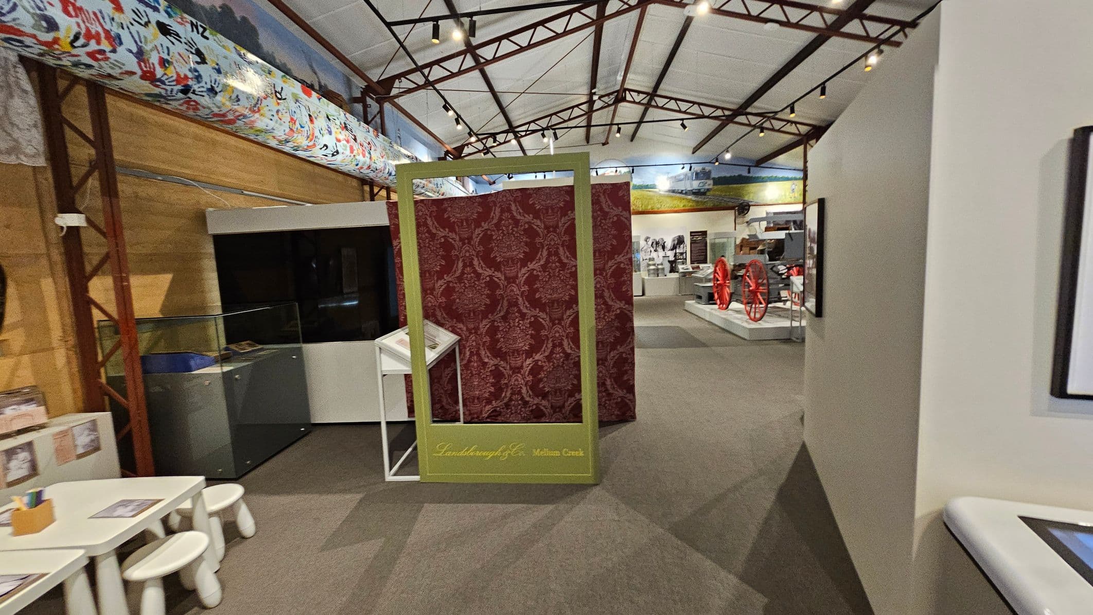 A museum interior with a high ceiling supported by metal beams. In the centre, a decorative red panel with a gold frame stands, labelled "Landsborough & Co. Melton Creek." To the left, a small table with stools and a display case. On the right, a wall with framed pictures. In the background, exhibits include red wagon wheels and historical artefacts. The floor is carpeted in a neutral tone.