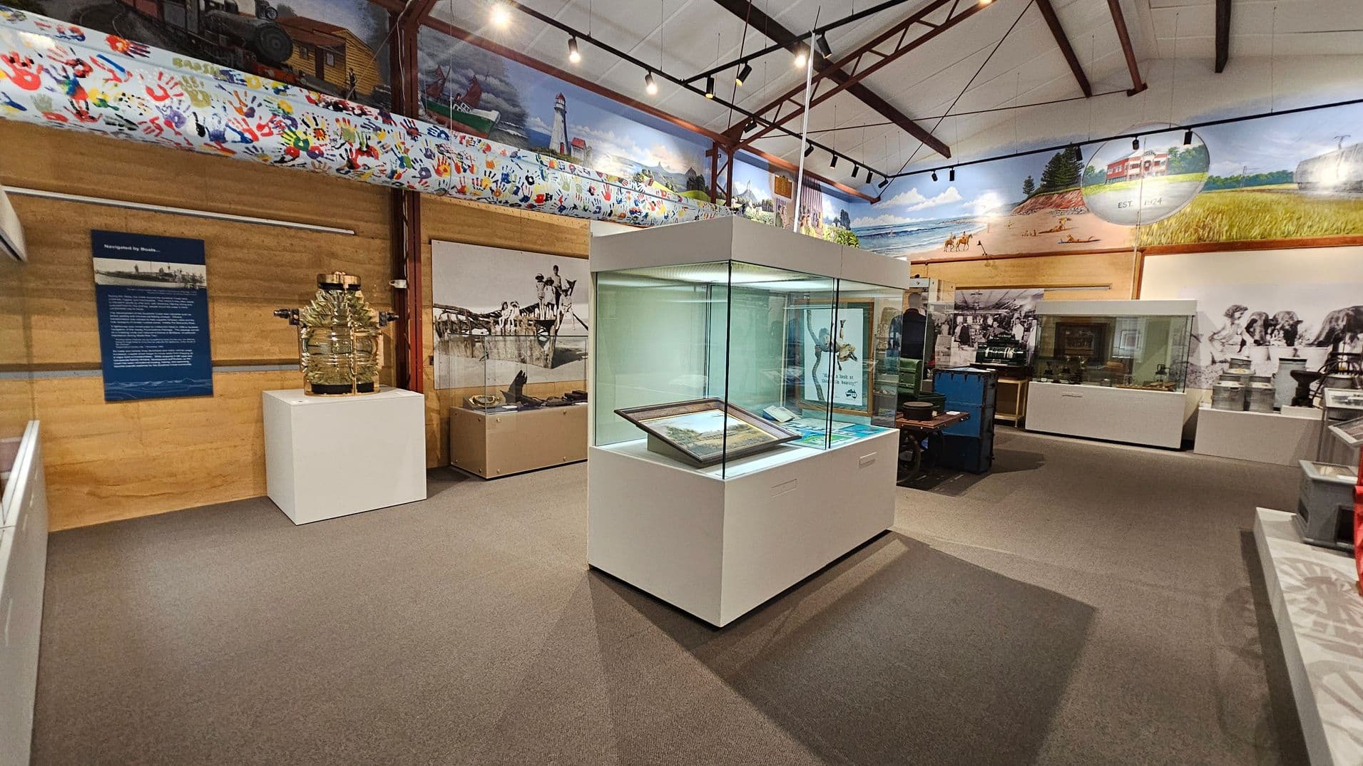 A museum interior with a central glass display case containing framed items. To the left, a large glass object on a white pedestal and a blue information panel are visible. The walls are adorned with murals depicting historical scenes and colourful handprints. The ceiling features exposed beams and track lighting. The floor is carpeted in grey. In the background, more display cases and historical photographs are present.