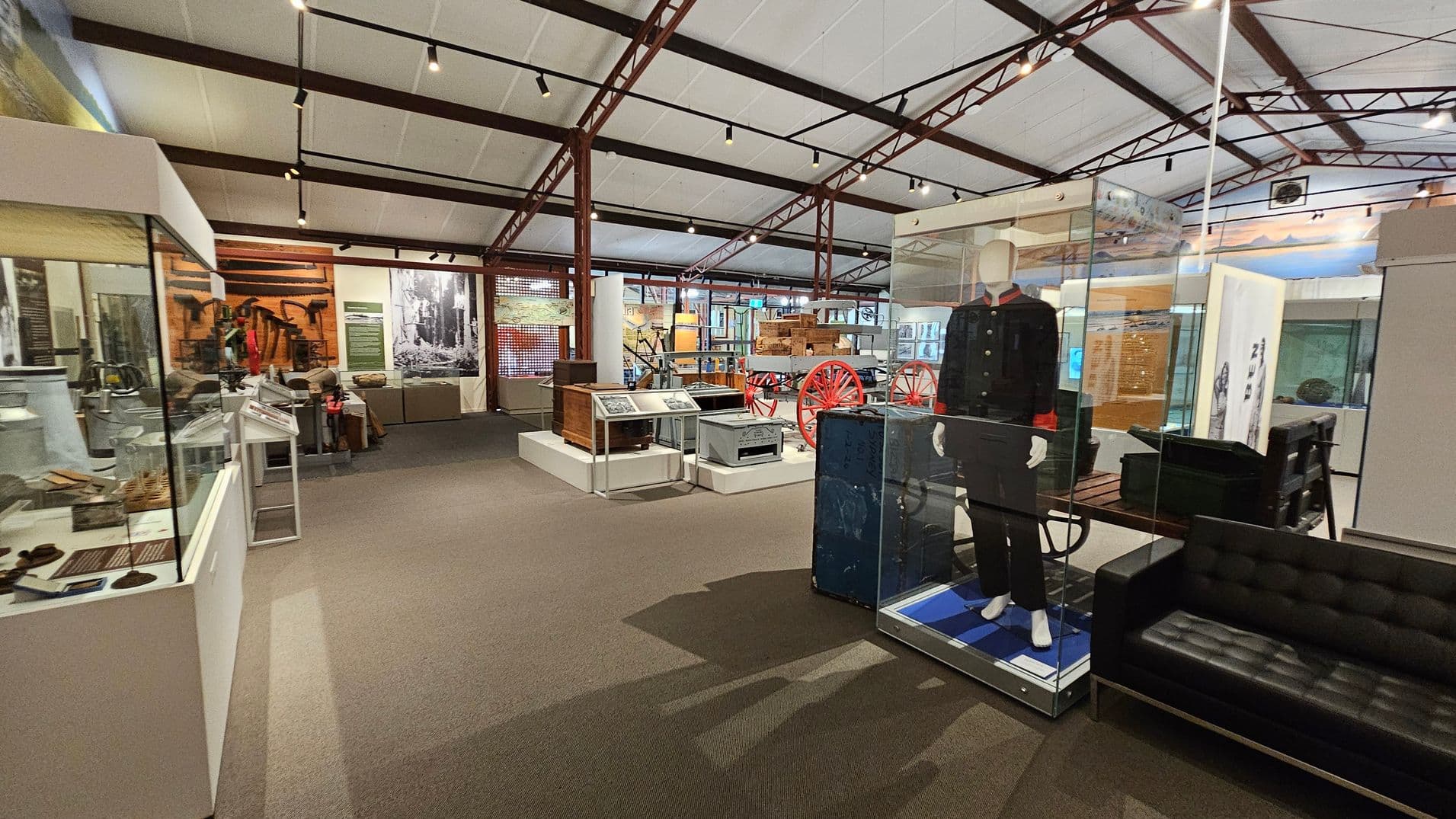 A museum interior with various exhibits. On the left, there are glass display cases containing artefacts. In the centre, a mannequin dressed in a historical uniform is encased in glass. Behind it, a red-wheeled cart is visible. The right side features a black leather sofa. The ceiling has exposed beams and track lighting, and the floor is carpeted. In the background, there are more displays and large black-and-white photographs on the walls.