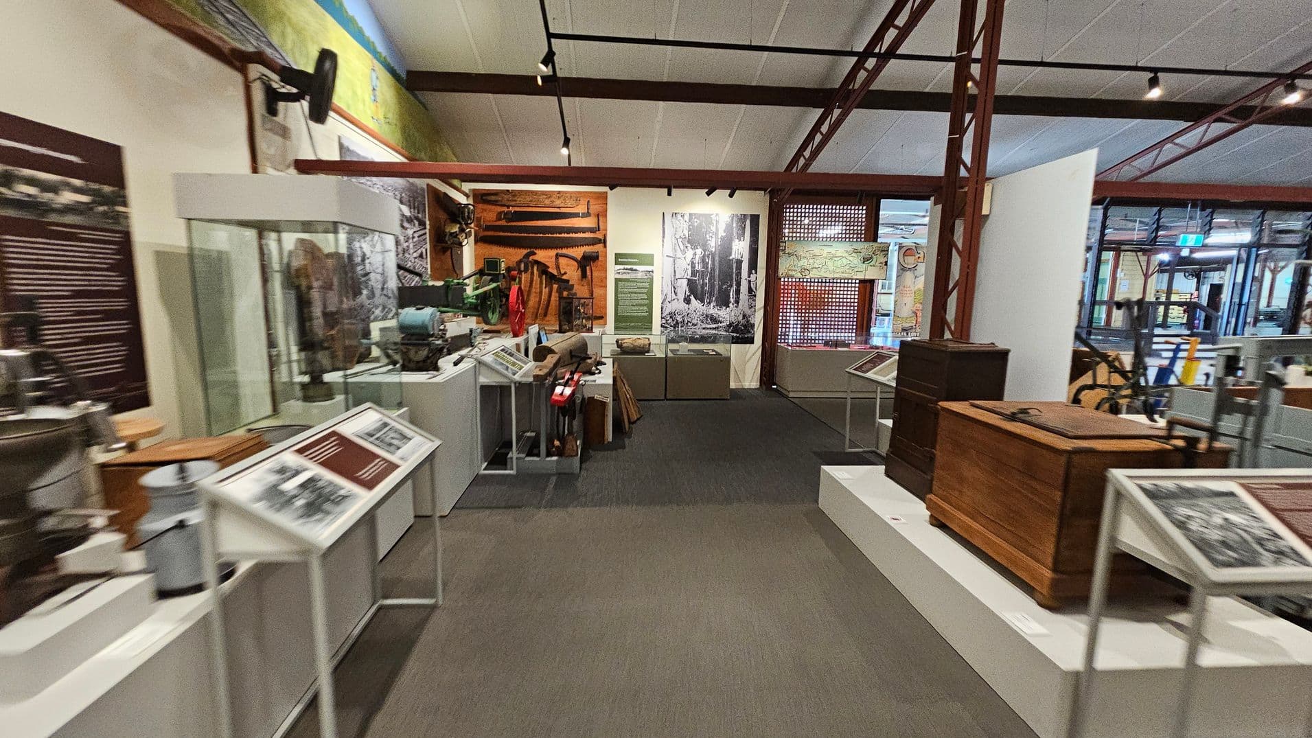 A museum exhibit with various historical artefacts. On the left, there are display cases with tools and informational panels. In the centre, a large black-and-white photograph is mounted on the wall, with more tools and equipment displayed below. On the right, wooden chests and additional artefacts are showcased. The floor is carpeted, and the ceiling has exposed beams with track lighting.