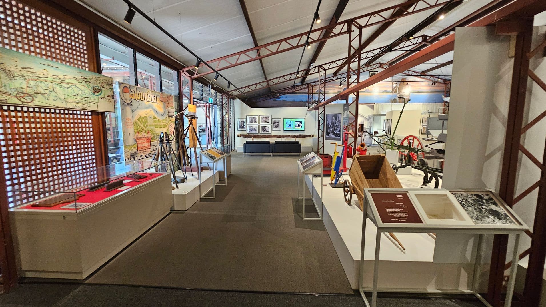 A museum exhibit with a high ceiling supported by metal beams. On the left, there's a display case with a red interior and a long object inside, alongside a wall with a large map and the word "Caloundra." In the centre, there are several tripods and information stands. On the right, a wooden wheelbarrow and other historical tools are displayed. The background features framed pictures and a screen above a black sofa. The floor is carpeted.