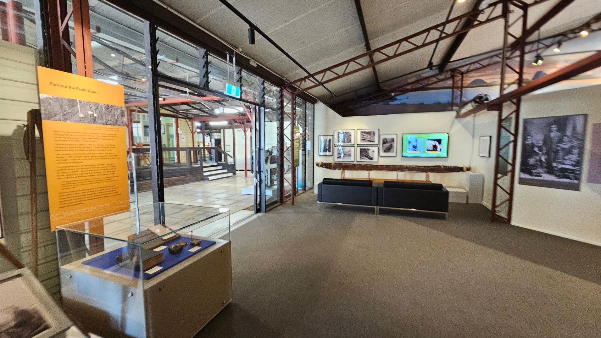 A museum interior with a glass display case on the left containing artefacts. An orange informational panel is nearby. The centre features a seating area with a black bench. On the right wall, there are framed photographs and a digital screen. The ceiling has exposed metal beams, and the floor is carpeted. In the background, a glass wall with a door leads to another section of the museum, visible through the glass.
