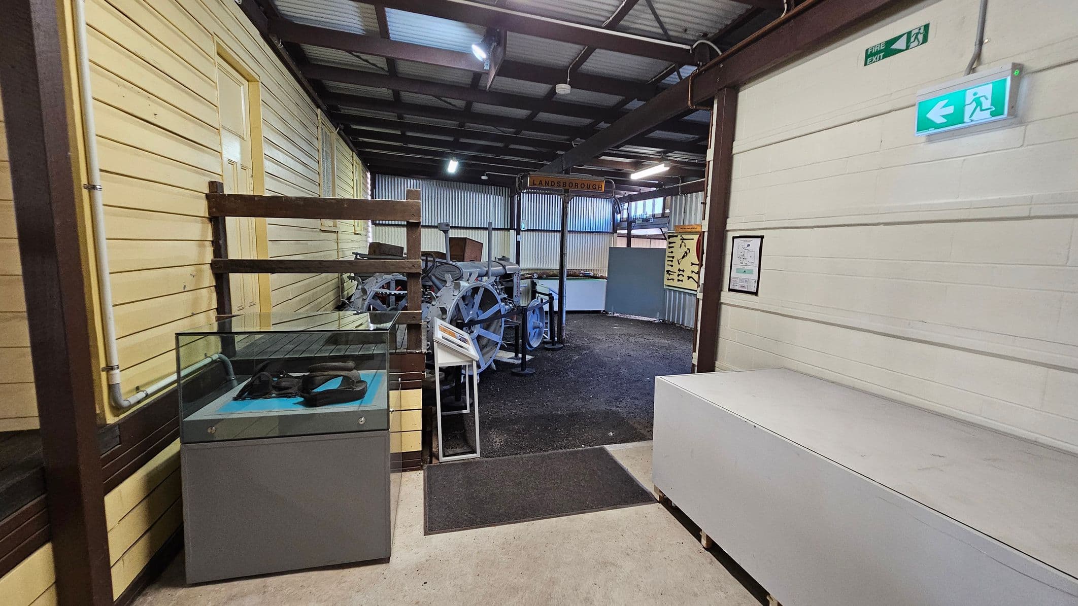 A room with a concrete floor and a corrugated metal ceiling. On the left, there's a wooden ramp with a "MACHINERY" sign. In the centre, a doorway leads to a space with machinery and display cases. On the right, a white brick wall features a small table with stools, colouring sheets, and markers. A shelf holds various items, including a green bag. A fire extinguisher is mounted on the wall near the doorway.