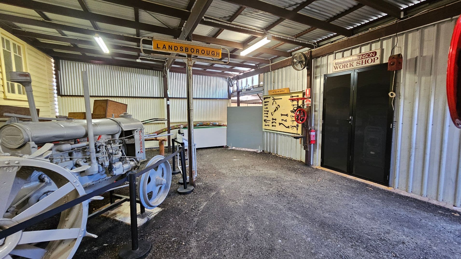 Inside a workshop with a corrugated metal ceiling and walls. On the left, there's a vintage Fordson tractor with large metal wheels. A sign above reads "LANDSBOROUGH." On the right, a black door labelled "WORK SHOP" is flanked by a red valve wheel and a fire extinguisher. A display board with various tools is mounted on the wall. The floor is gravel, and fluorescent lights hang from the ceiling.