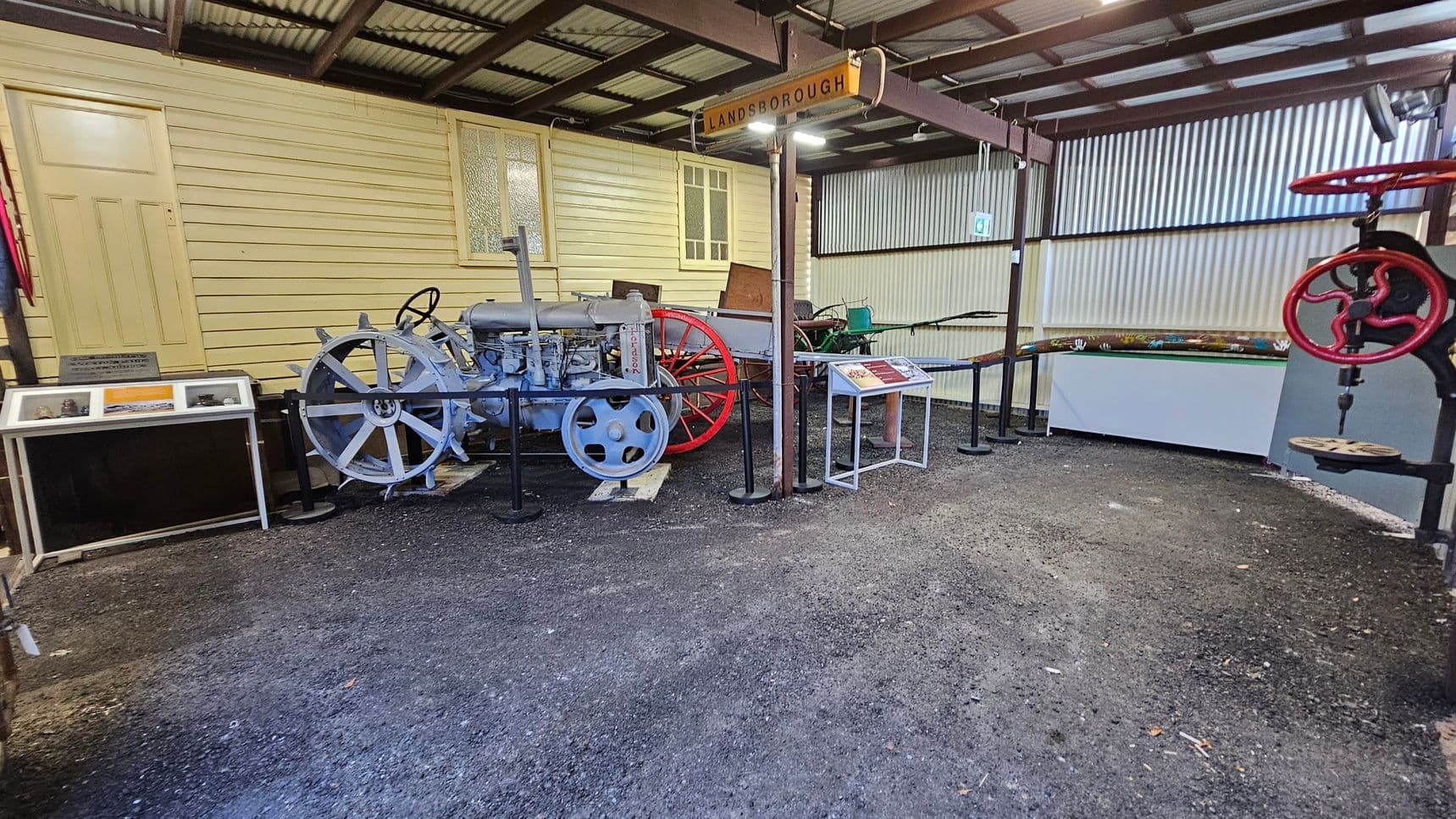 A museum exhibit featuring vintage machinery. On the left, there's a light blue tractor with large spoked wheels, displayed in front of a yellow wooden wall with a door and window. A sign above reads "LANDSBOROUGH." To the right, there's a red wheel mechanism and informational plaques. The floor is gravel, and the ceiling is corrugated metal.