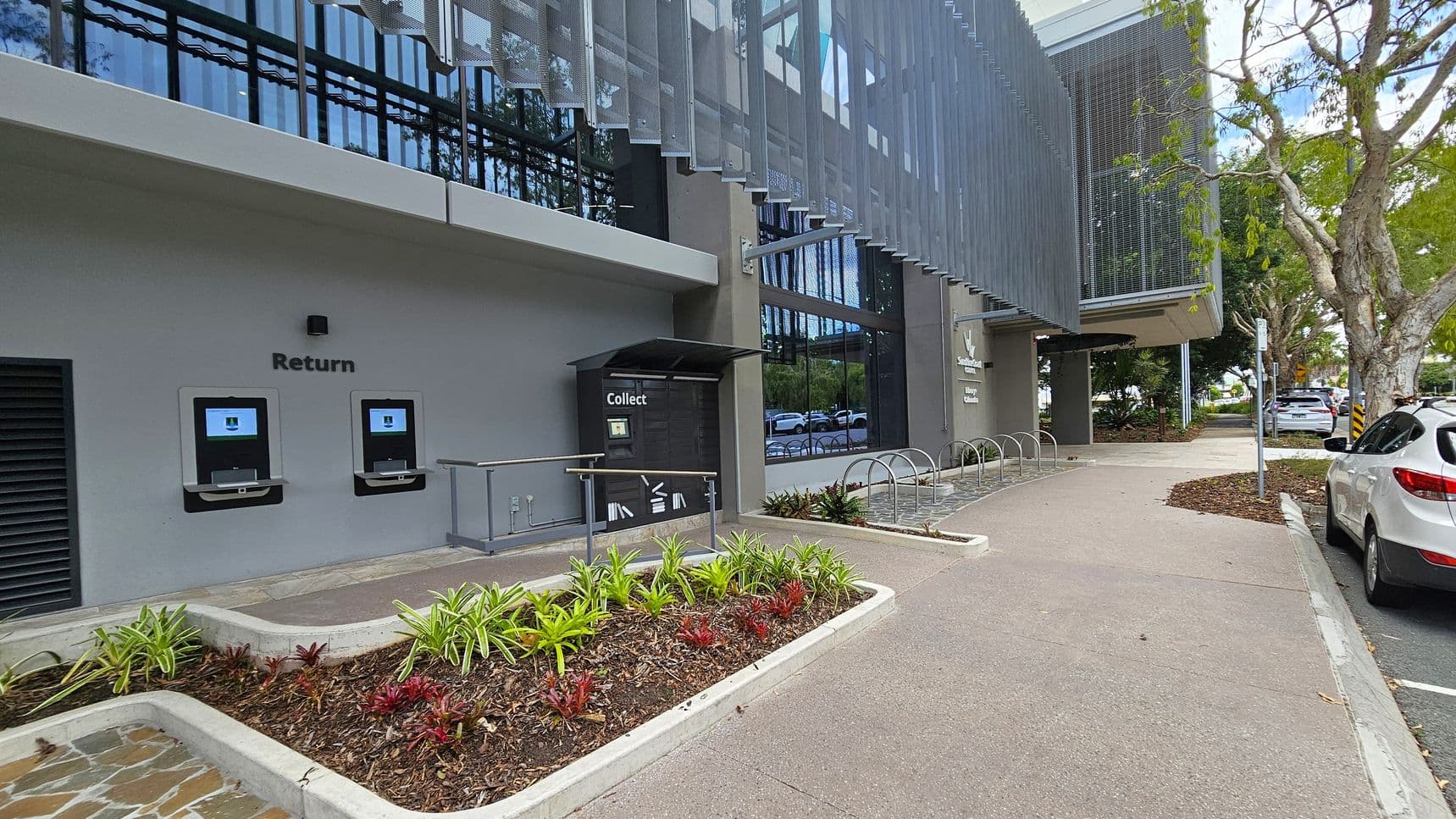 On the left, there are two automated return machines embedded in a grey wall, labelled "Return". Below them is a small garden bed with various plants. In the centre, a "Collect" locker is situated under a small awning. The building features large glass windows with a metal mesh awning above. On the right, a paved path leads past a bicycle rack and parked cars. Trees line the street, providing shade.