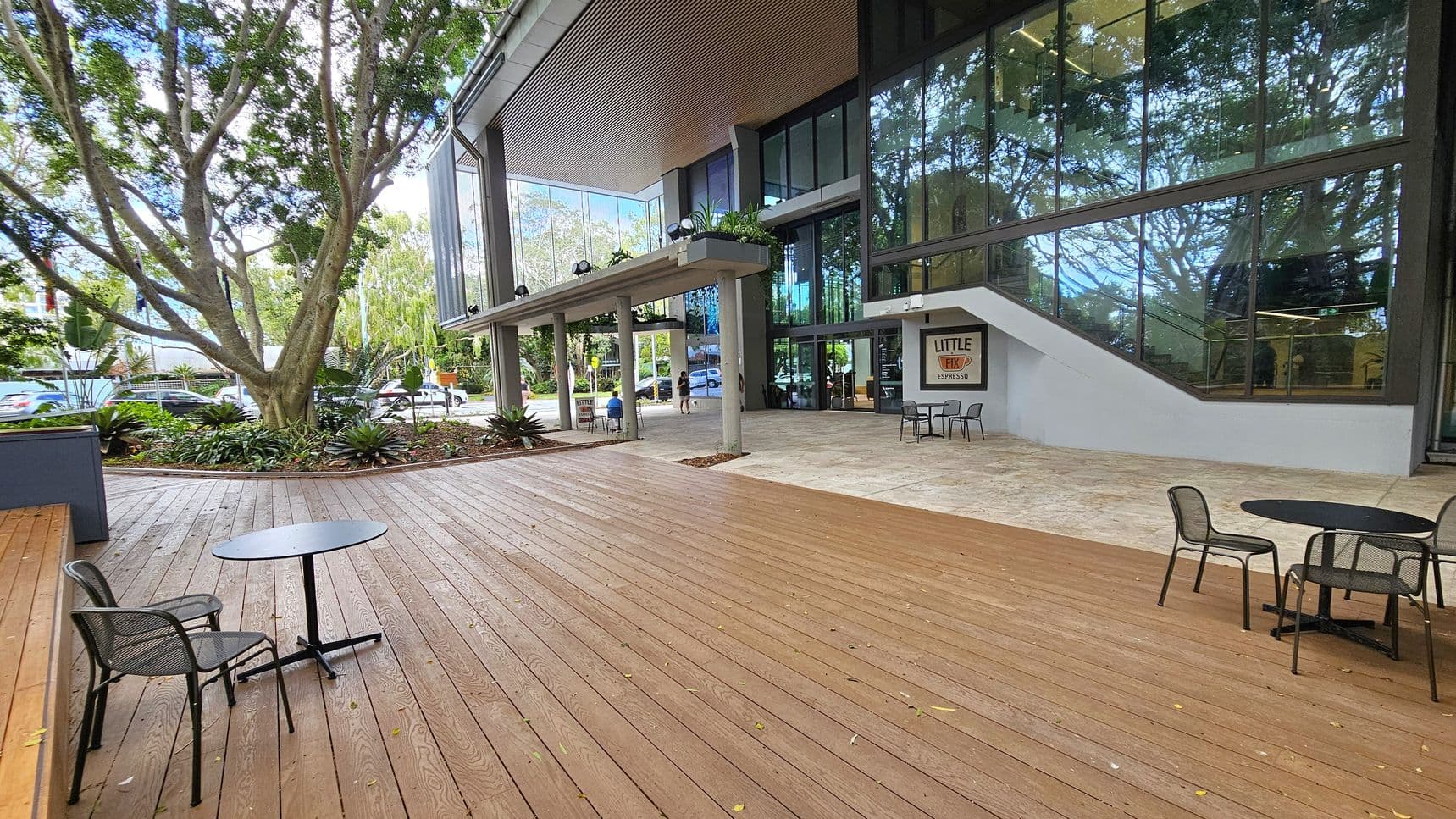 A spacious outdoor area with wooden decking on the left, featuring a round table and chairs. A large tree with a thick trunk and lush foliage is surrounded by plants. On the right, a modern building with large glass windows and a sign for "Little Fix Espresso" near the entrance. The building has a covered walkway supported by columns. The ground transitions from wood to stone tiles near the building.