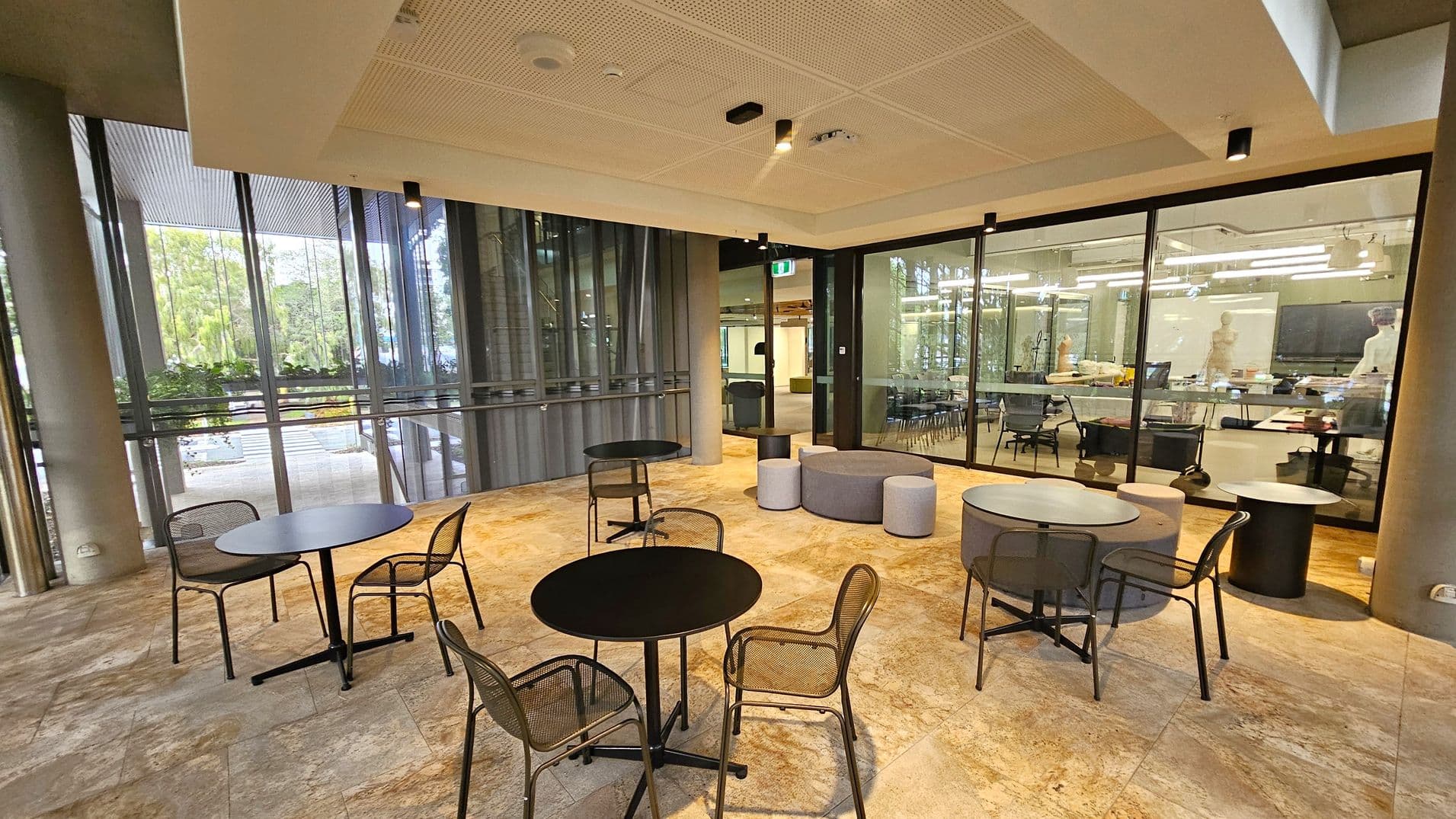 A modern indoor seating area with round black tables and mesh chairs on a stone-patterned floor. On the right, there are grey and purple cushioned stools. The background features large glass windows and doors, revealing a well-lit room with tables, chairs, and a whiteboard. The ceiling is light-coloured with recessed lighting and ventilation fixtures. Outside the windows, greenery is visible.