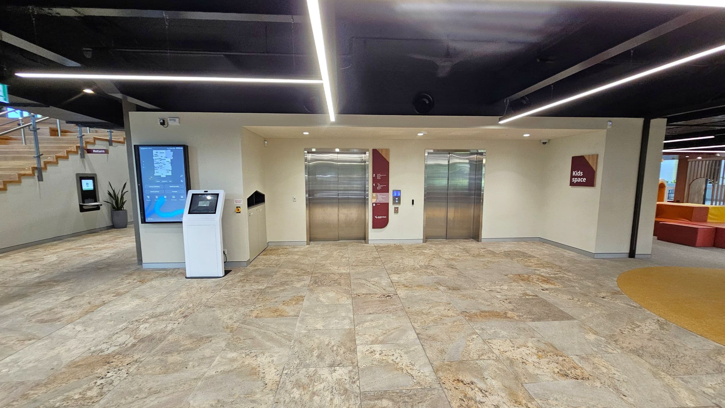 Two lifts with metallic doors are centred on a light-coloured wall. To the left, a digital directory and a white kiosk stand near a staircase with wooden steps and a potted plant. On the right, a sign reads "Kids space" above a colourful seating area. The floor is tiled with a mix of beige and green hues, and the ceiling features black panels with linear lighting.