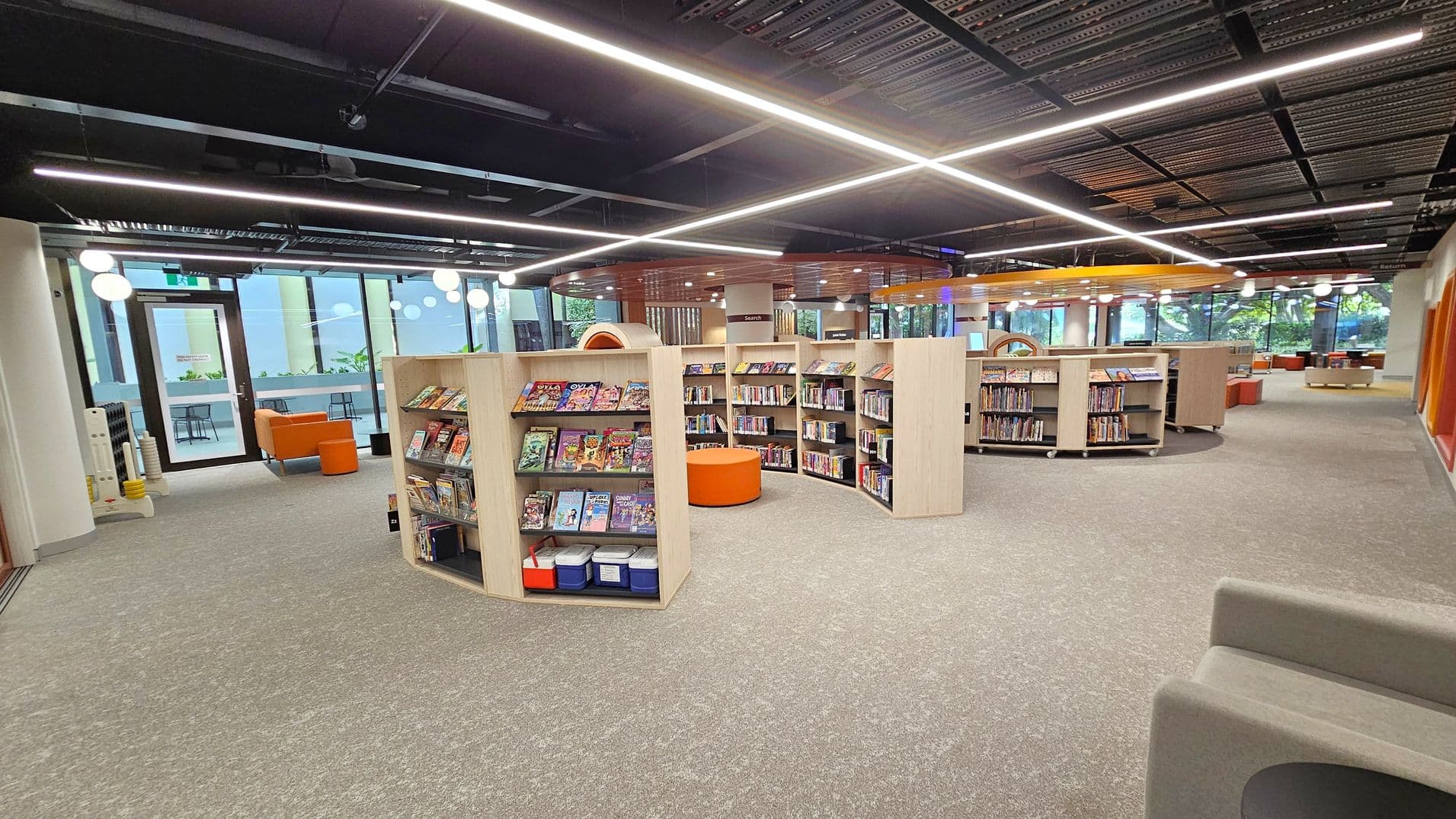 A modern library interior with light wood bookshelves arranged in a semi-circular pattern, filled with books and comics. In the centre, there is an orange circular seating area. The floor is carpeted in grey. Large windows on the left and background allow natural light in, with a view of greenery outside. The ceiling is black with linear lighting fixtures. Orange and grey seating is visible near the windows, and there are round white ceiling lights throughout.