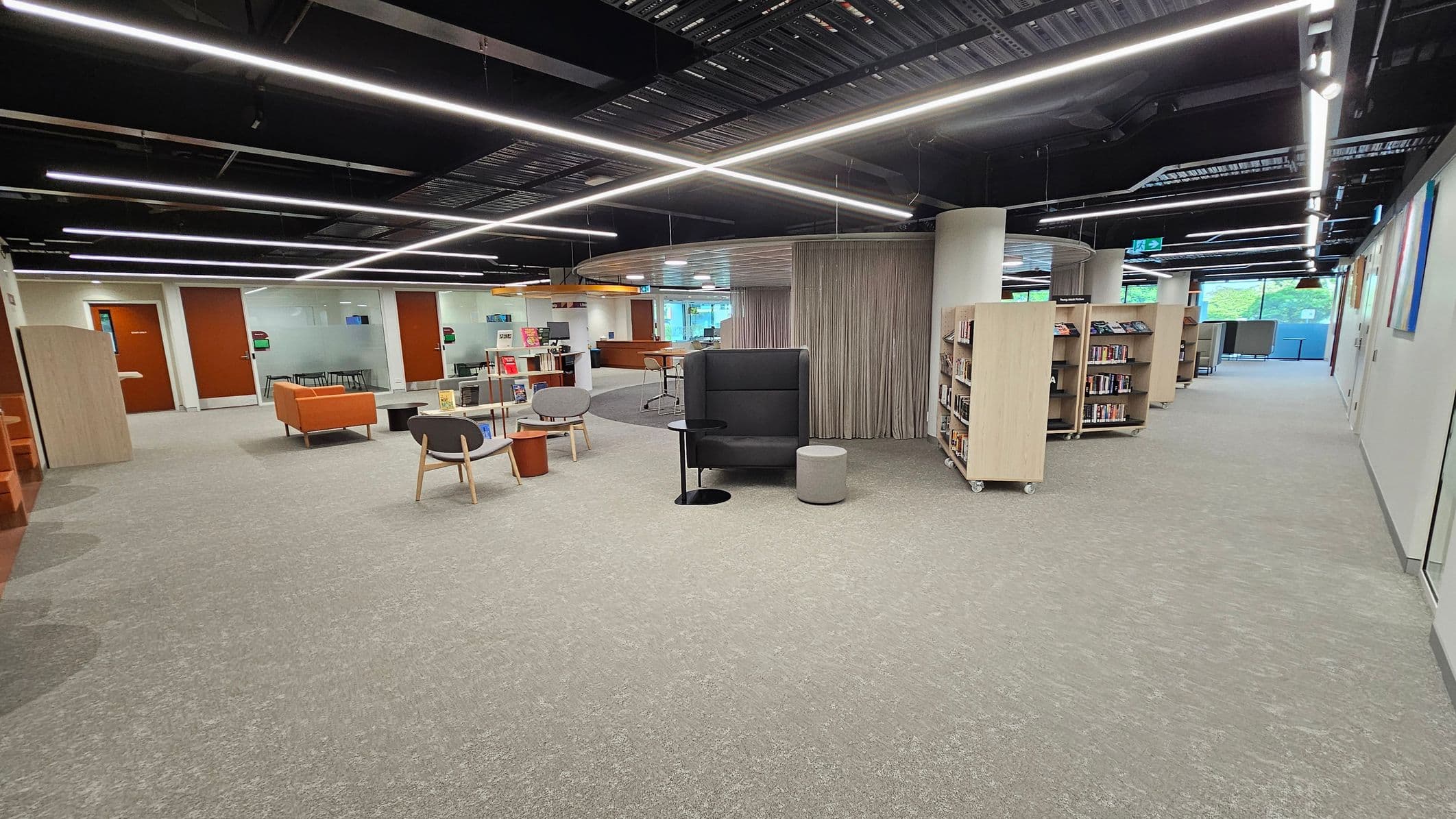 A modern library interior with a light green carpet. On the left, there are orange sofas and grey chairs around small tables. In the centre, a high-backed black chair with a small round table and stool. On the right, bookshelves on wheels line the wall. The ceiling features exposed beams and linear lights. In the background, glass walls and doors lead to additional rooms, with a view of greenery through large windows.