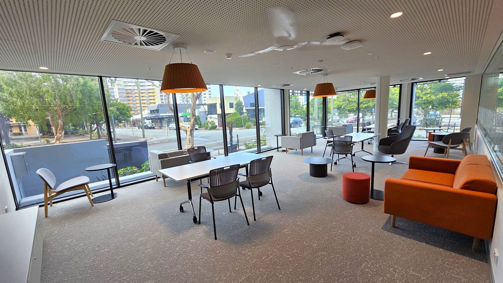 A modern office lounge with large floor-to-ceiling windows on the left and right, offering views of trees and buildings outside. The room features a mix of seating, including grey chairs, black mesh chairs, and an orange sofa. White tables are arranged in the centre, with small round tables scattered around. The ceiling has recessed lighting, air vents, and large pendant lights. The floor is carpeted in grey.