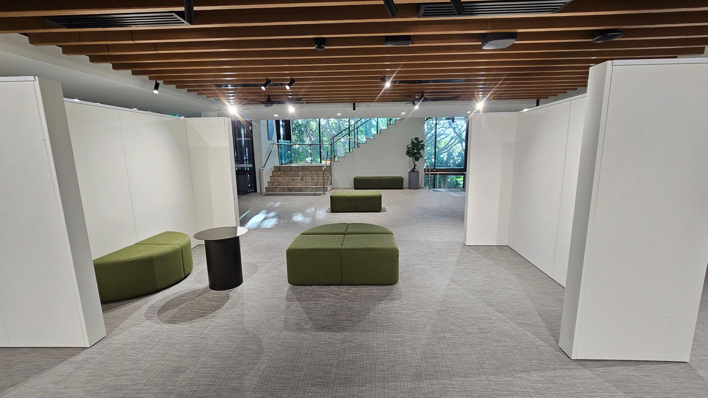 A modern interior space with grey carpeted flooring and a wooden slatted ceiling. In the centre, there are green cushioned benches and a small round black table. On the left, a curved green bench is positioned against a white partition wall. At the back, a staircase with glass railings leads upwards, flanked by large windows showing greenery outside. A potted plant is placed near the stairs. The lighting is provided by spotlights on the ceiling.