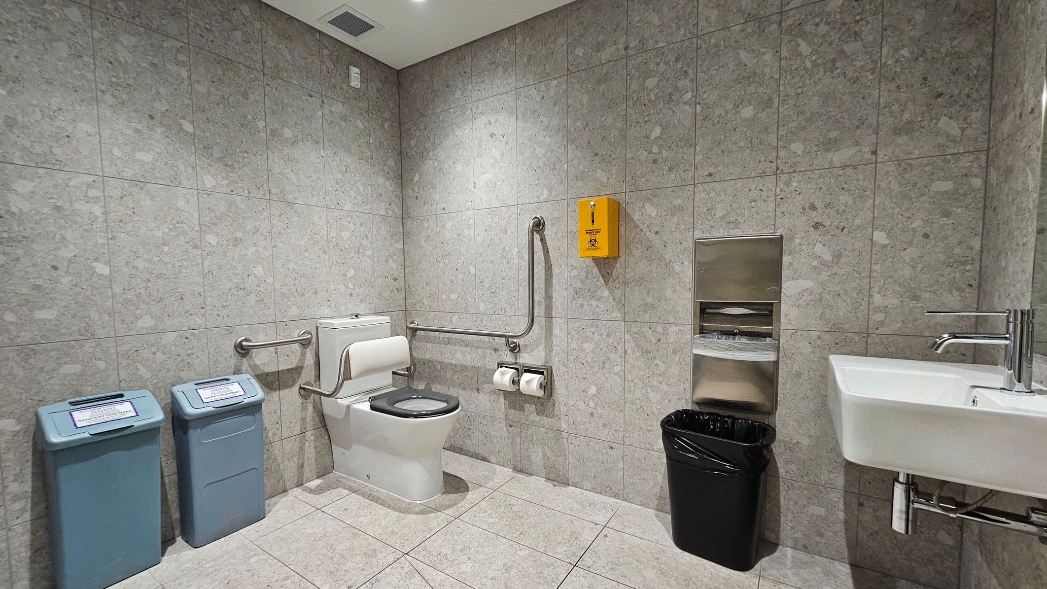 A public restroom with grey-green tiled walls and floor. On the left, there are two blue waste bins with labels. In the centre, a toilet with a black seat is equipped with support rails and two toilet paper rolls. Above the toilet, a yellow biohazard disposal box is mounted. On the right, a white sink with a silver tap is next to a black waste bin. Above the bin, a metal paper towel dispenser is attached to the wall. The ceiling is white with a vent and recessed lighting.