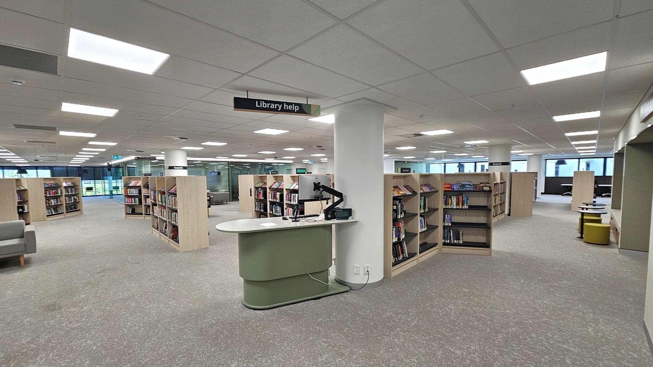 A modern library interior with a grey and green carpeted floor. In the centre, a curved help desk with a computer is positioned near a white column. Bookshelves filled with books are arranged in rows throughout the space. Overhead, a sign reads "Library help." The ceiling is fitted with rectangular fluorescent lights. Large windows in the background allow natural light to enter, and there are seating areas with grey sofas and small tables.
