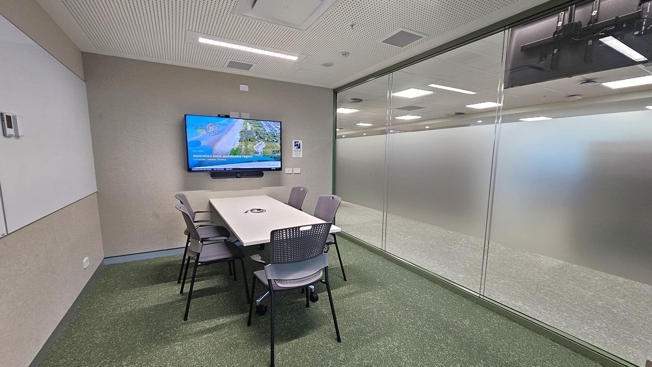 A small meeting room with a rectangular white table surrounded by six grey chairs. On the left wall, a large screen displays an image and text. The right side features a glass wall with a frosted lower section, revealing a view of an adjacent room with ceiling lights. The floor is carpeted in green, and the ceiling is white with recessed lighting and ventilation panels.