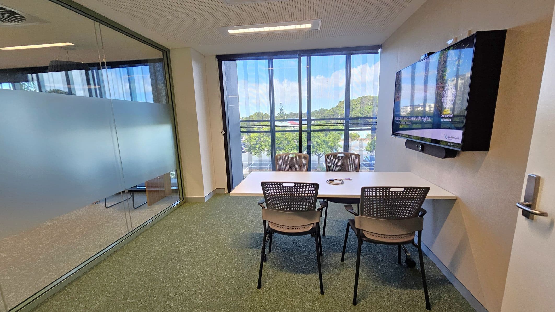 A small meeting room with a glass wall on the left, partially frosted for privacy. In the centre, a rectangular table with three chairs. On the right wall, a large flat-screen monitor is mounted. The room has a green carpet and a white ceiling with recessed lighting. The far wall features large windows with vertical blinds, offering a view of trees and a parking area outside. A door with a handle is visible on the right.