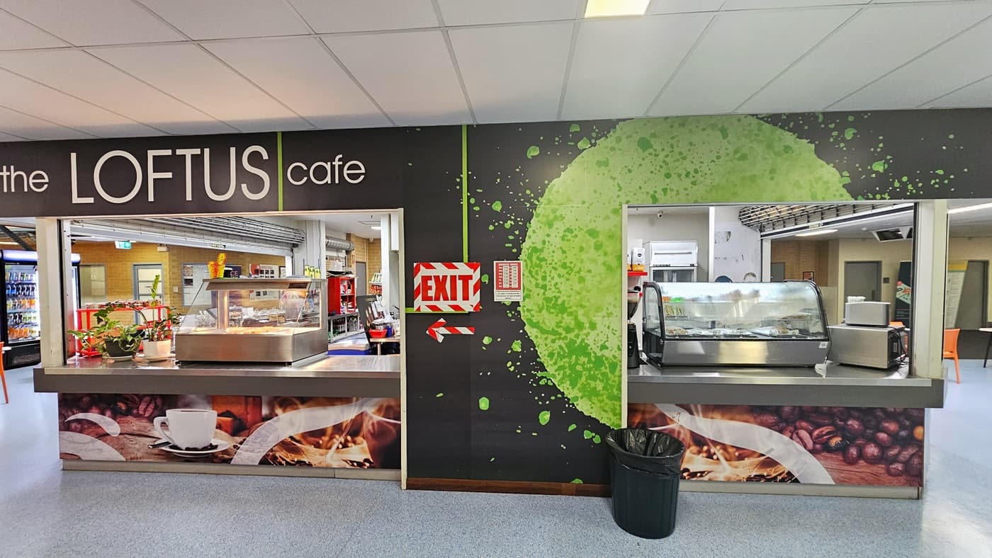 A cafe adorned with coffee granule imagery, featuring a menu posted in the corner. A food display rack is positioned on top of the counter, with a black rubbish bin nearby. On the left, black and orange plastic chairs and a black leather couch are arranged for seating. A ramp leading to the change facility is also visible. To the right, a lobby leads to the reception, with movable plastic chairs and tables in place. The floor has blue vinyl, and the ceiling is entirely white.