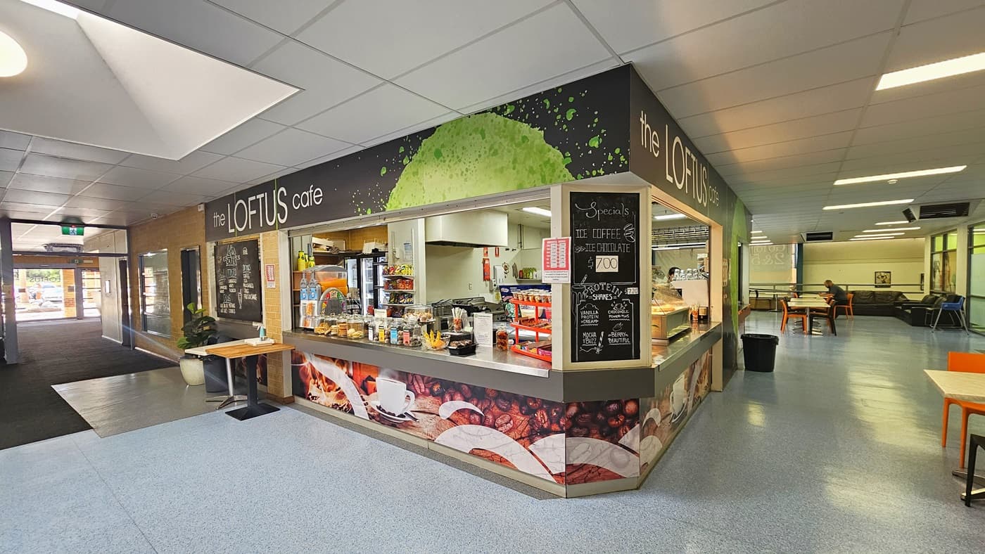 A cafe adorned with coffee bean imagery, identified by the sign "The Loftus Cafe." The counter is stocked with food items for sale, and a menu is posted in the right corner. A wooden table near the counter holds a hand sanitizer, alongside a rubbish bin and a potted ornamental plant. On the right, black and orange plastic chairs are visible, along with another rubbish bin. The floor is covered with blue vinyl, and the ceiling is entirely white.