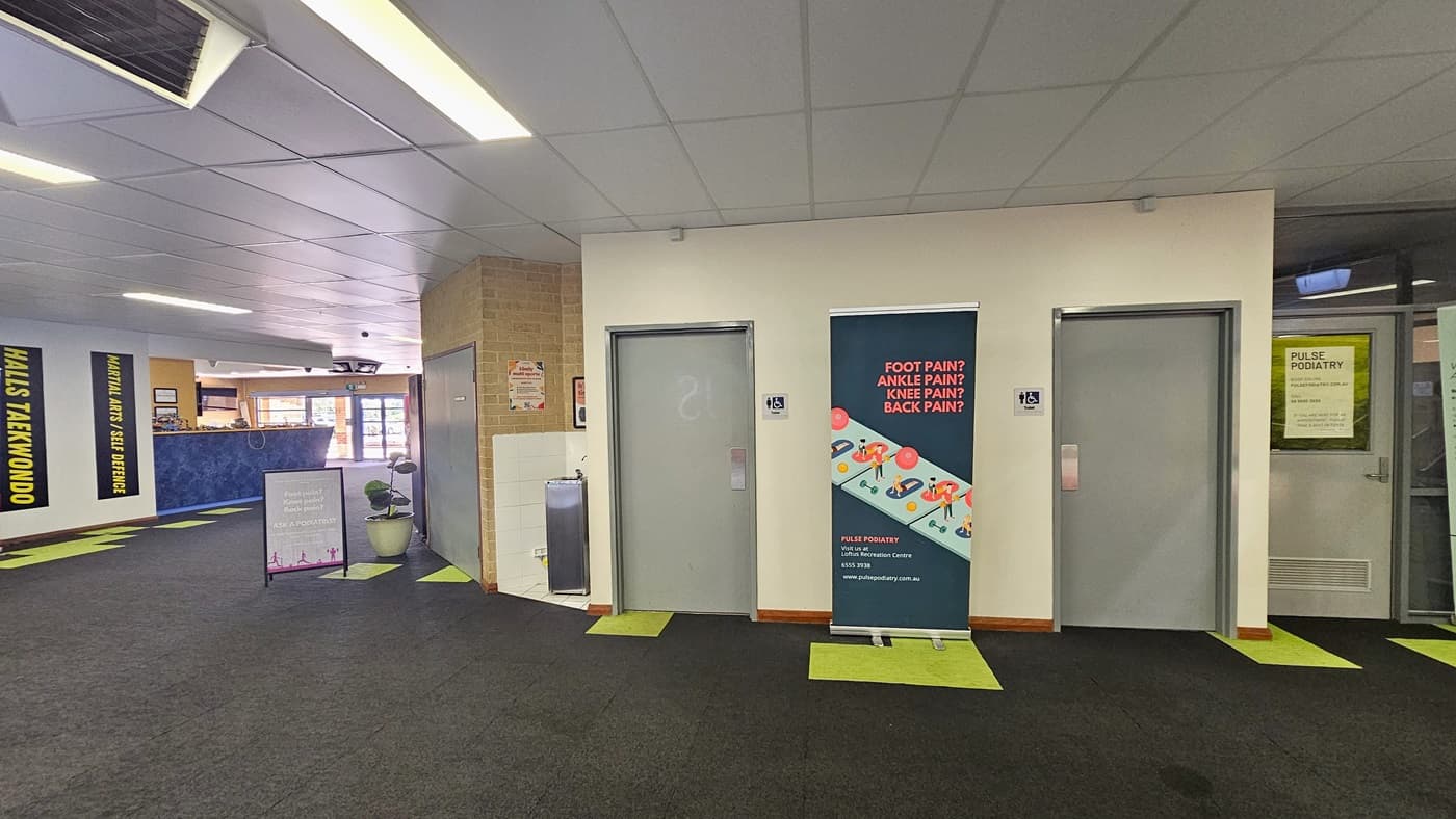 Accessible bathrooms, marked by two grey doors. A large advertising banner is between the doors, with another banner and a potted plant on the left. The reception desk is on the left.  To the right is another door with a poster. To the right is another door with a banner. The ceiling is white with air conditioning vents and fluorescent lights, and the floor is grey with neon green accents.