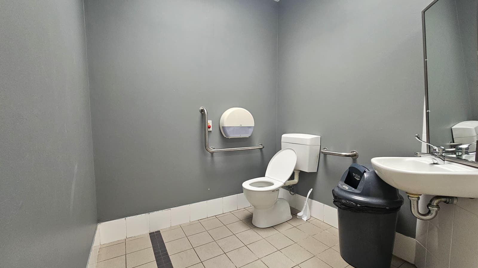 Within the accessible bathroom, a toilet is positioned in the left corner, equipped with handrails. A wall-mounted tissue dispenser is on the left. To the right, a washbasin and a mirror are affixed to the wall, with a black rubbish bin underneath. The floor is covered with white tiles, and the wall is a plain grey.