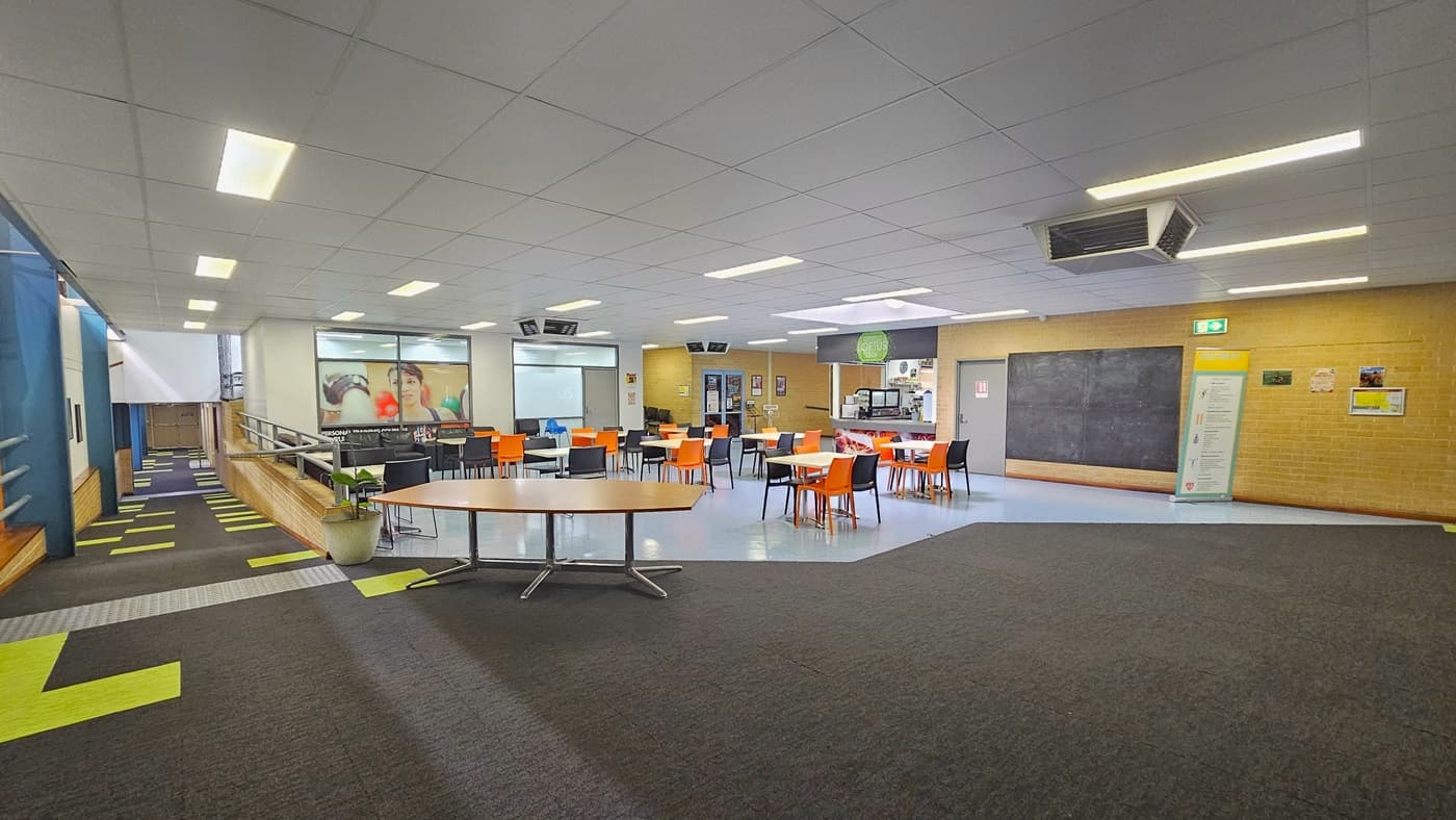 Black and orange movable plastic chairs and tables are arranged. To the left, there's a ramp leading to the change room with truncated domes and handrails. Large banners are displayed on the left brick wall, including a fire exit sign. On the far right is the cafe counter. The floor on the right has grey tiles accented with neon green tiles, while the floor on the left has blue vinyl. The ceiling is white with large air conditioning vents.