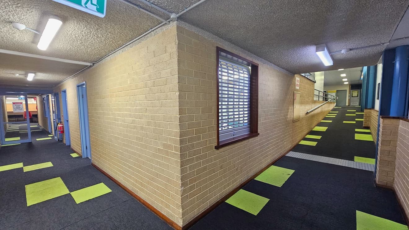 To the right is a ramp equipped with handrails and truncated domes. The middle wall comprises a cream brick wall with a window and posters. The right wall is accented with blue. To the left, there is a glass door with grey frames. The ceiling features a rough grey design with fluorescent lights. A green exit sign is also visible. The floor is covered with grey tiles, accented with neon green.