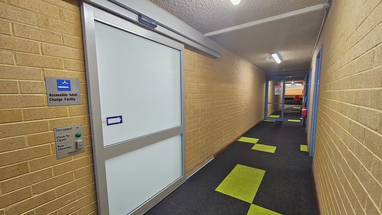 The corridor has glass doors with grey frames. The entrance to the accessible adult change facility is through a white sliding door marked by an arrow pointing right. Signage is mounted on the walls. Both sides have brown brick walls, and the ceiling is white with fluorescent lights. The floor is grey with neon green accents. A fire extinguisher is mounted on the right wall, and grey glass doors are also visible.