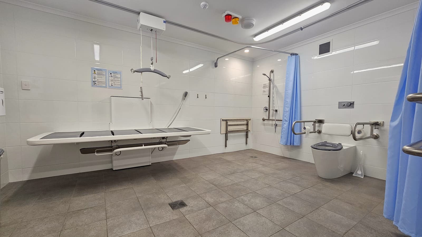 On the left is an adult change table with a hoist. Next to it is a shower with handrails and a blue curtain. A foldable bench is mounted to the wall. In the middle is a toilet bowl equipped with handrails and a tissue dispenser. The left wall has another handrail with a shower curtain. The bathroom is spacious with grey floor tiles and white wall tiles. A banner that bears instructions is posted on the left wall.