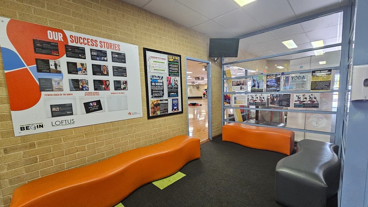 In the Group Fitness Room there is seating arranged with orange and black cushioned chairs. The cream brick wall is adorned with success story posters and features a flat-screen TV. Inside, the fitness room showcases timber walls with white ceilings, and additional posters adorn the right wall.