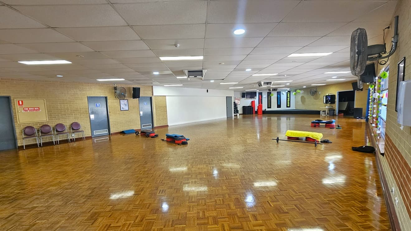 The fitness room features polished timber flooring. Fitness equipment is stored on the far left. An accessible bathroom, marked for gender access, is on the left, and brown chairs are arranged in front. Wall fans are installed on the surrounding brick walls. Large windows on the far right feature some decorations and offer a view outside. The ceiling is white with aircon vents.