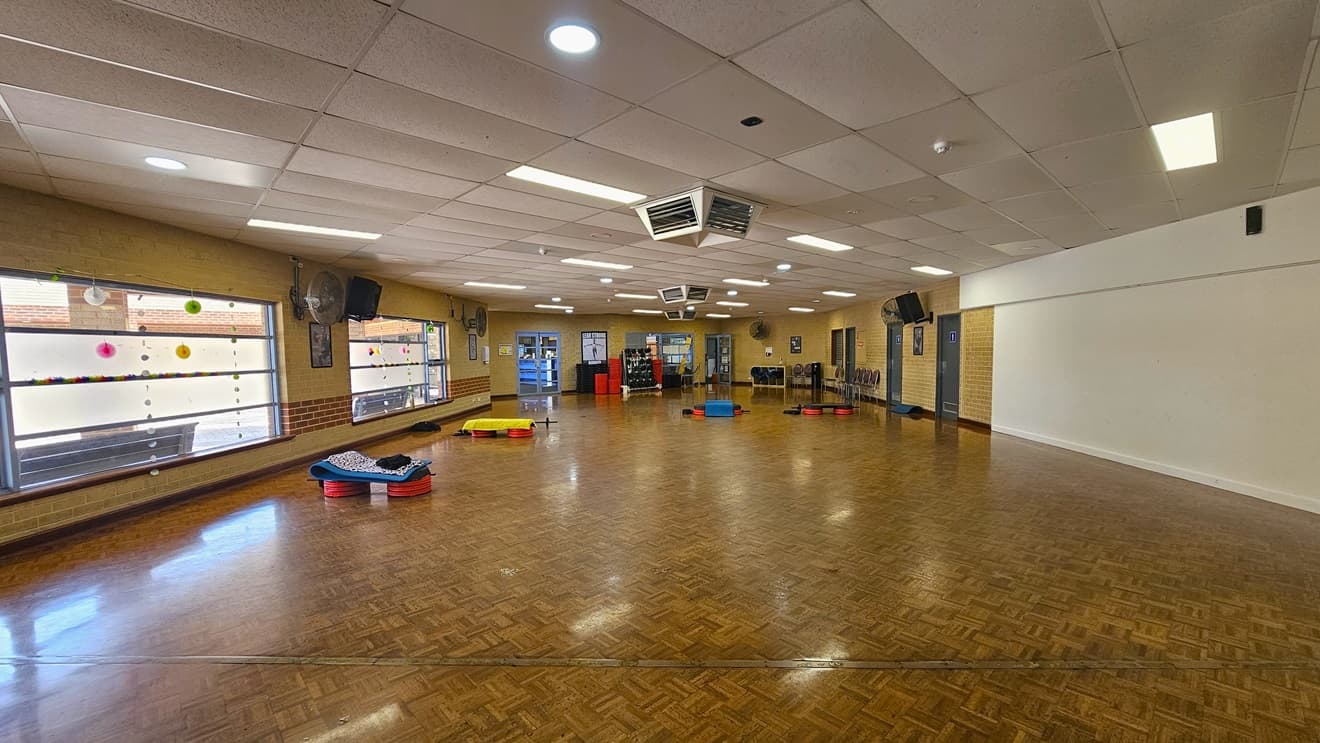 The fitness room has polished timber flooring, with fitness equipment stored at the far end along with yoga mats. An accessible bathroom, marked for gender access, is on the right, and brown chairs are arranged in front. Wall fans are installed on the surrounding brick walls. Large windows on the left feature some decorations and offer a view outside. The ceiling is white with aircon vents.