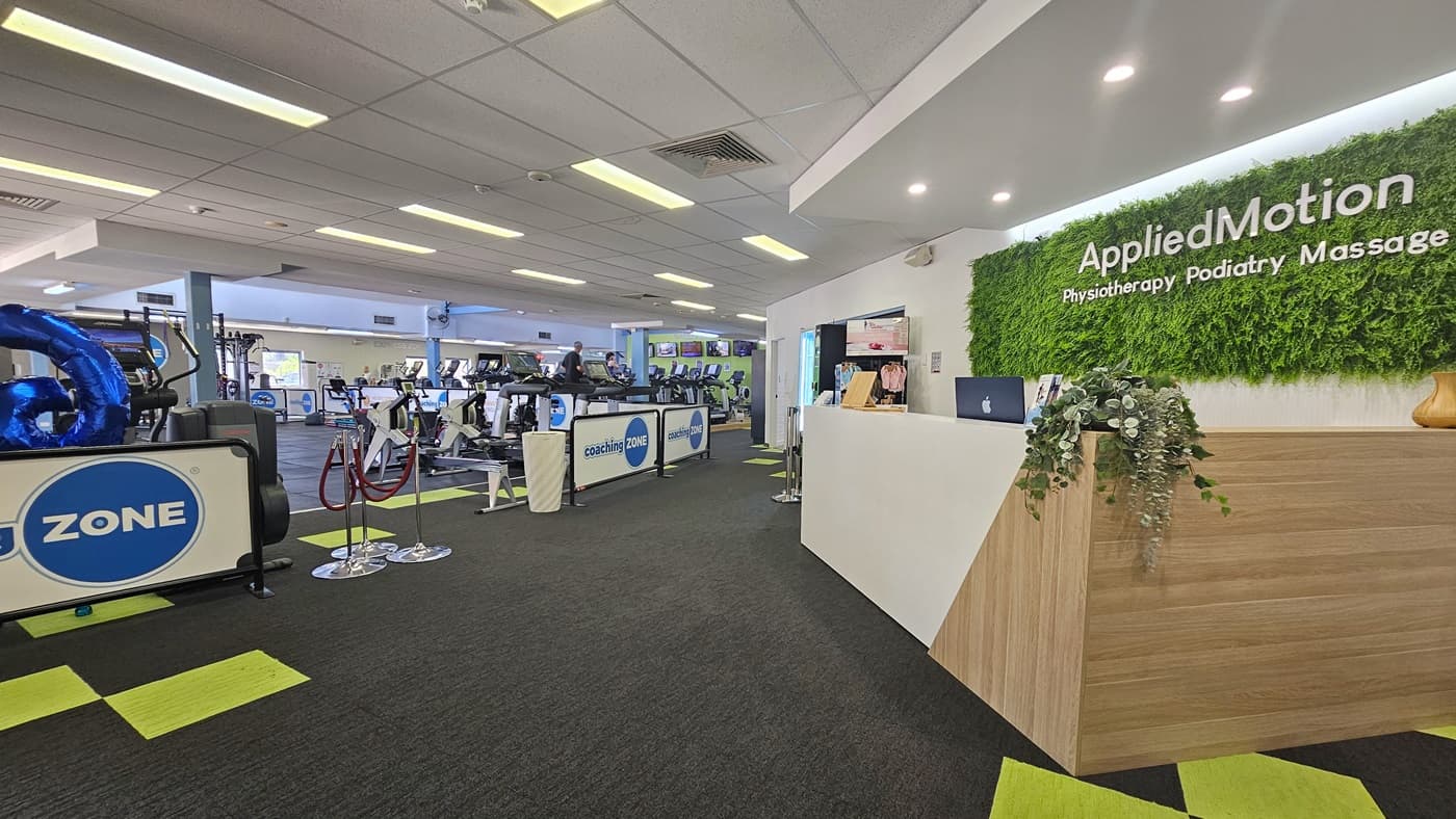 The reception desk for Applied Motion, enclosed with glass. Banners are attached to the wall. To the left is a cream and brown brick wall with a large informational banner. To the right is another brick wall, where a vending machine for cold drinks is visible. An orange cushioned chair is against the wall, accompanied by a wall clock and a fire hose reel overhead. The ceiling is white, and the floor has grey tiles with neon green accents. A potted ornamental plant is placed near the signage.