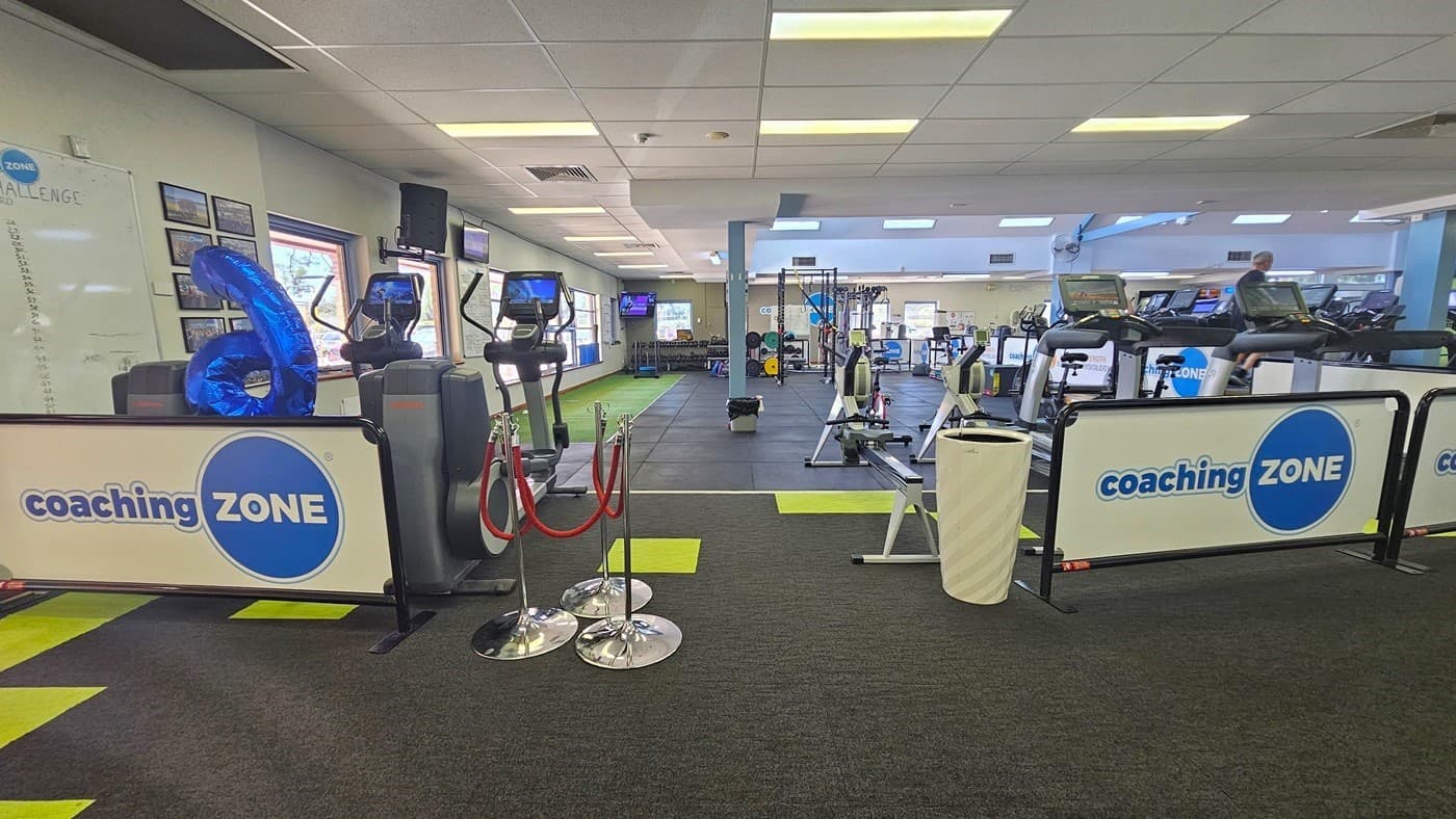 "Coaching sessions” barricade boards line the front entrance, with a central white rubbish bin. On the left, a large blue number balloon and cardio machines are present, alongside a rope bollard. To the right, rowing and treadmill machines are visible. The floor has neon green carpet tiles outside and grey rubber tiles inside. Posters and a whiteboard adorn the left wall, while the ceiling is entirely white.