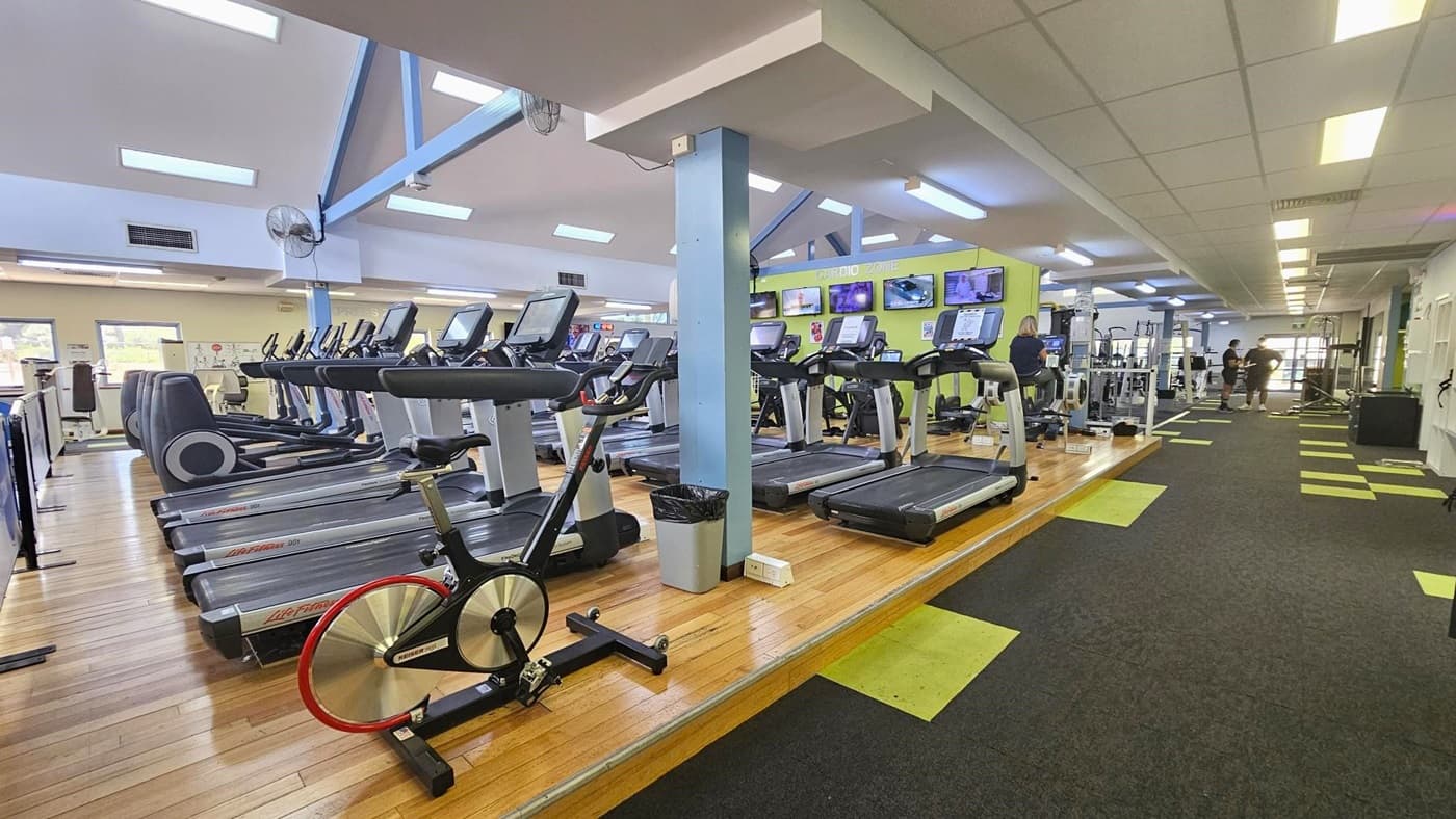 The gym's left section has a timber floor with treadmills and a spin bike. Moving in, there are additional machines. Flat-screen TVs are mounted on the green wall in the middle. To the right, an area with grey carpet and neon green tiles is present. The ceiling is white with fluorescent lights.