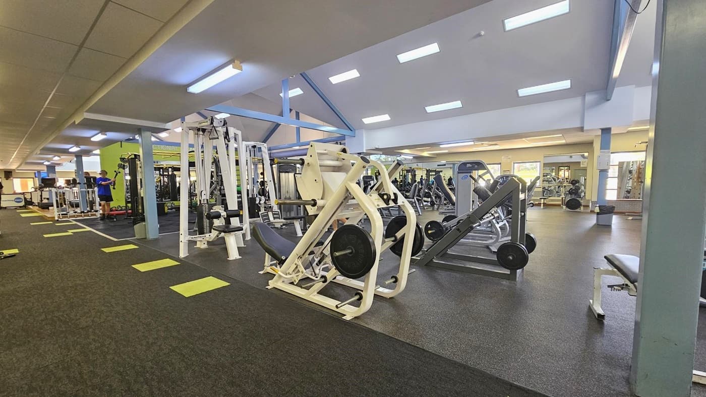 On the right, there are bench press machines, incline bench press, smith machines, and lat pull-down machines. Moving further in, additional machines are present. The floor is tiled in grey with neon green accents. The ceiling is white with fluorescent lights. Gym Goers are also visible.