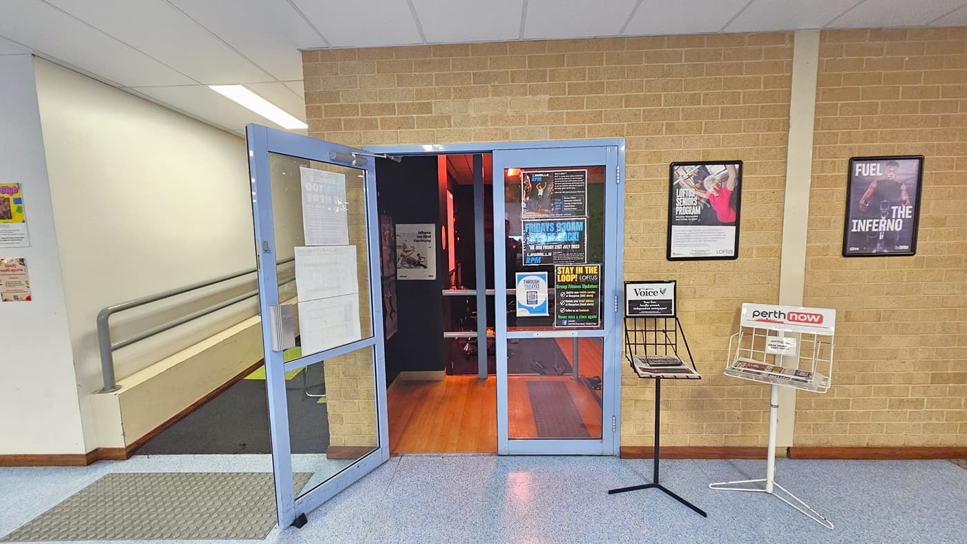 On the left, is a ramp entry equipped with handrails and truncated domes. In the middle, there is a manual glass door that opens outward. Newspaper stands are positioned on the right, and posters are affixed to the brick wall, including the glass door. The floor outside is equipped with grey tiles, and inside, it is made of timber. The interior is a dimly lit area.