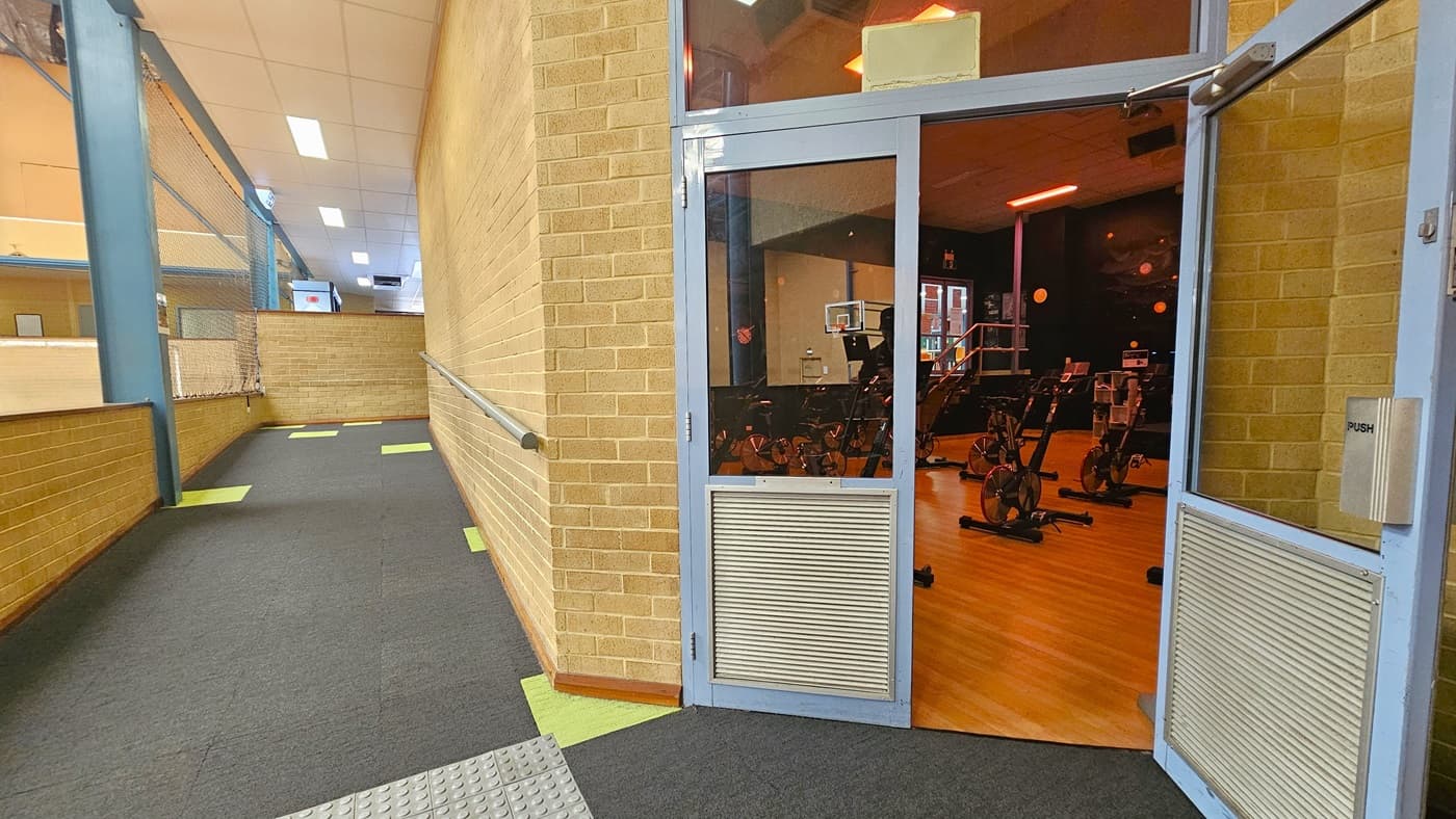 On the left, there is a ramp entry equipped with truncated domes. To the right is a glass door that opens outward. Inside, the floor is made of timber, and stationary bikes are arranged. The space inside is dimly lit. The wall outside is made of brown bricks, and the ceiling is white with fluorescent lights. The floor has grey tiles with neon green accents.